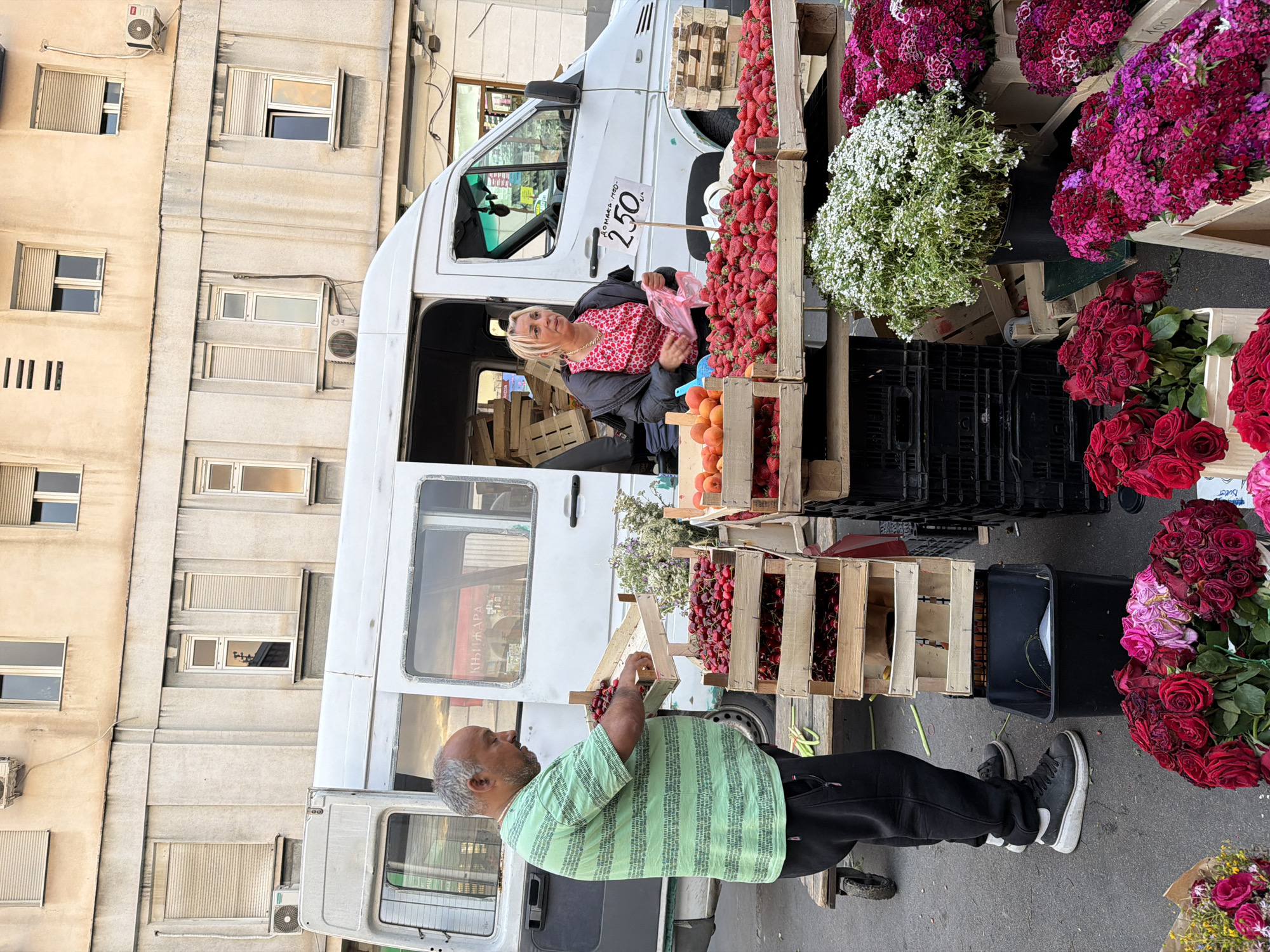 The image depicts an outdoor market scene in Vracar Urban Municipality, Serbia. A white van serves as a stall, with a woman inside, possibly a vendor. The van displays produce: strawberries, oranges, and cherries are visible. A man in a green striped shirt is examining cherries. To the right, bouquets of flowers, primarily red and purple, are displayed. In the background, an apartment building is visible, indicating an urban setting.