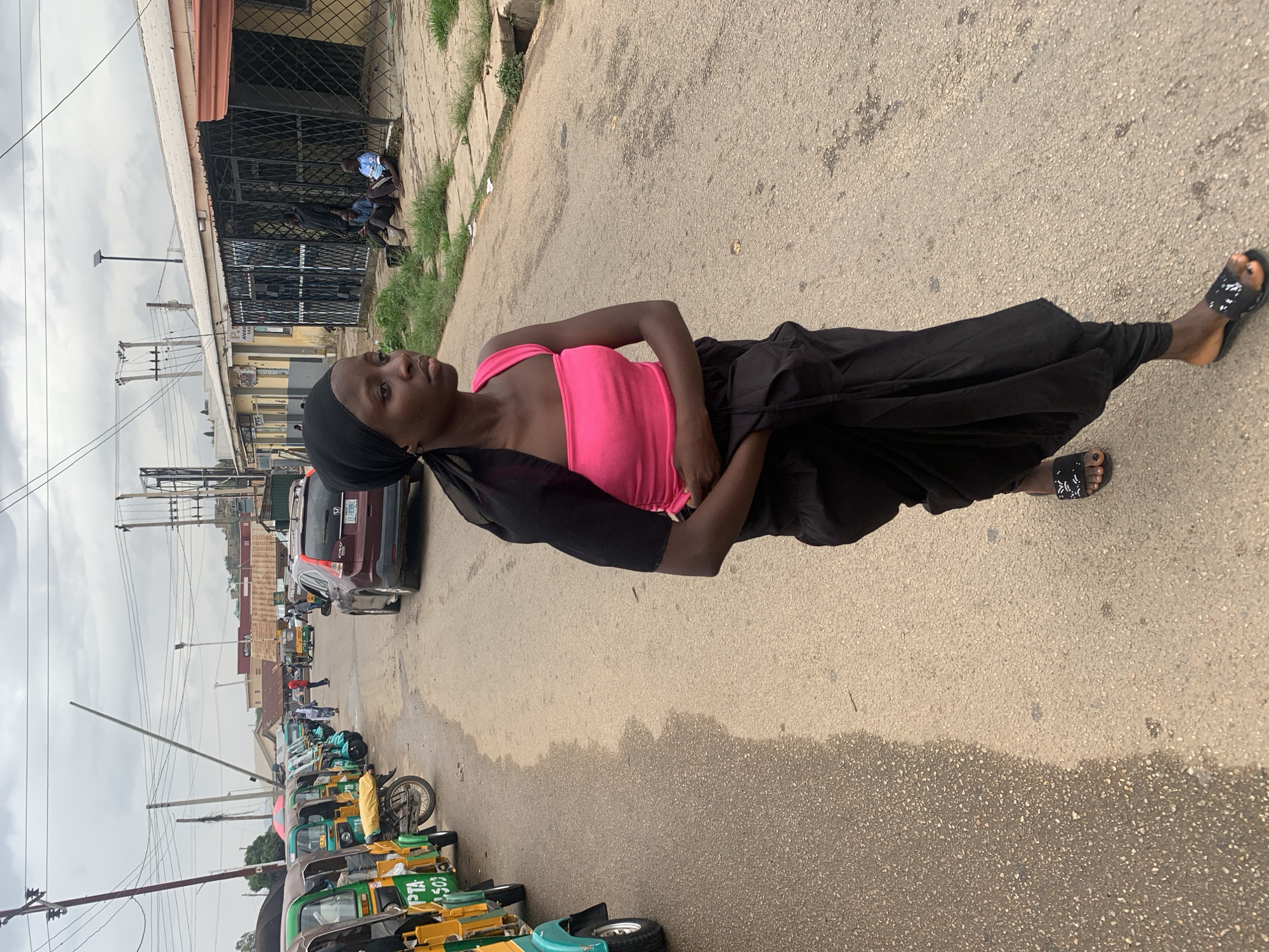 A young woman stands on the edge of a paved road, appearing to be in Jos, Nigeria. She is wearing a pink tank top, black head covering, and a long black skirt or pants. A black garment is draped over her shoulder. The road is bordered by a line of green and yellow tricycle taxis on one side. In the background, there are buildings with metal security gates, parked cars, and utility poles with numerous wires. The sky is overcast, and the road appears to be wet in places. There are several people visible in the distance beyond the cars and tricycles.