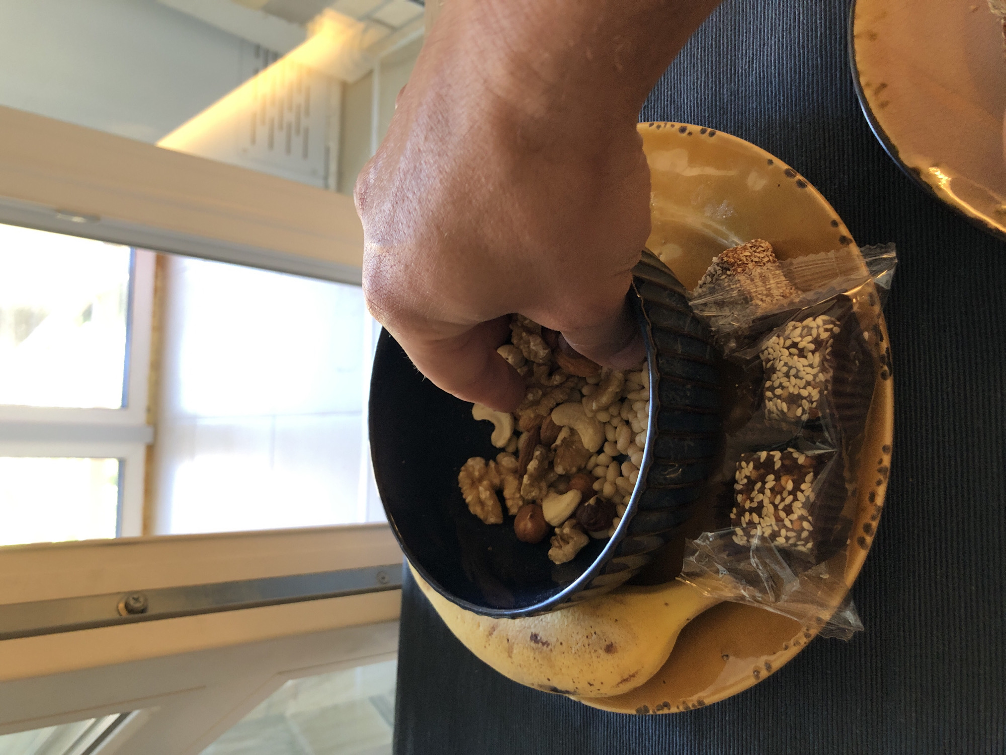 The image presents an indoor scene, likely set in Saint Petersburg, Russia, featuring a casual snack or meal setup by a window. In the foreground, a light-skinned adult hand, characterized by visible veins and skin texture, is reaching into a dark, fluted ceramic bowl filled with a variety of nuts and seeds. The bowl's contents include shelled walnuts, cashews, hazelnuts, pecans, and white pine nuts.
The bowl rests on a round, rustic-looking ceramic plate of an ochre-yellow color, distinguished by a subtly textured rim with dark, irregular speckles. Positioned on the left side of this plate is a ripe yellow banana, showing a few brown spots. To the right of the bowl, on the same plate, are two small, square-shaped dark confectioneries, each individually wrapped in clear plastic and generously topped with white sesame seeds. The plate and its contents are placed on a dark, horizontally ribbed placemat or tablecloth.
The background is dominated by a brightly lit window system with white frames, suggesting a multi-pane or modern window design. A slender, silver-colored metal strip with two visible fasteners runs vertically within one of the window frames. The outdoor view through the window is blurred but indicates a bright, daytime setting with hints of light-colored buildings or structures. Strong natural light floods the scene from the window, creating a warm ambiance and highlighting an angled interior wall or ceiling, possibly suggestive of a dormer window or an attic space.