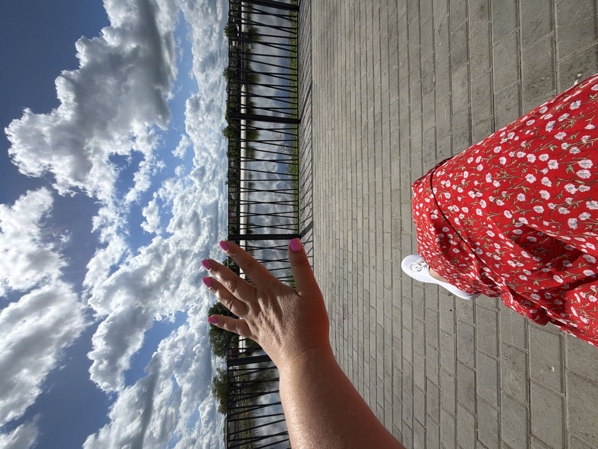 A first-person perspective photograph shows a person standing on a brick sidewalk alongside a black metal fence, likely in Leningradskaya Oblast, Russia, given the file name. The fence separates the sidewalk from a body of water that reflects the cloudy sky above. The person extends their right arm toward the sky; their hand has pink nail polish. The person wears a red dress with small white floral patterns and white sneakers. The sky is visible behind the fence, filled with fluffy white clouds against a blue sky. The ground is paved with gray bricks in a herringbone pattern.