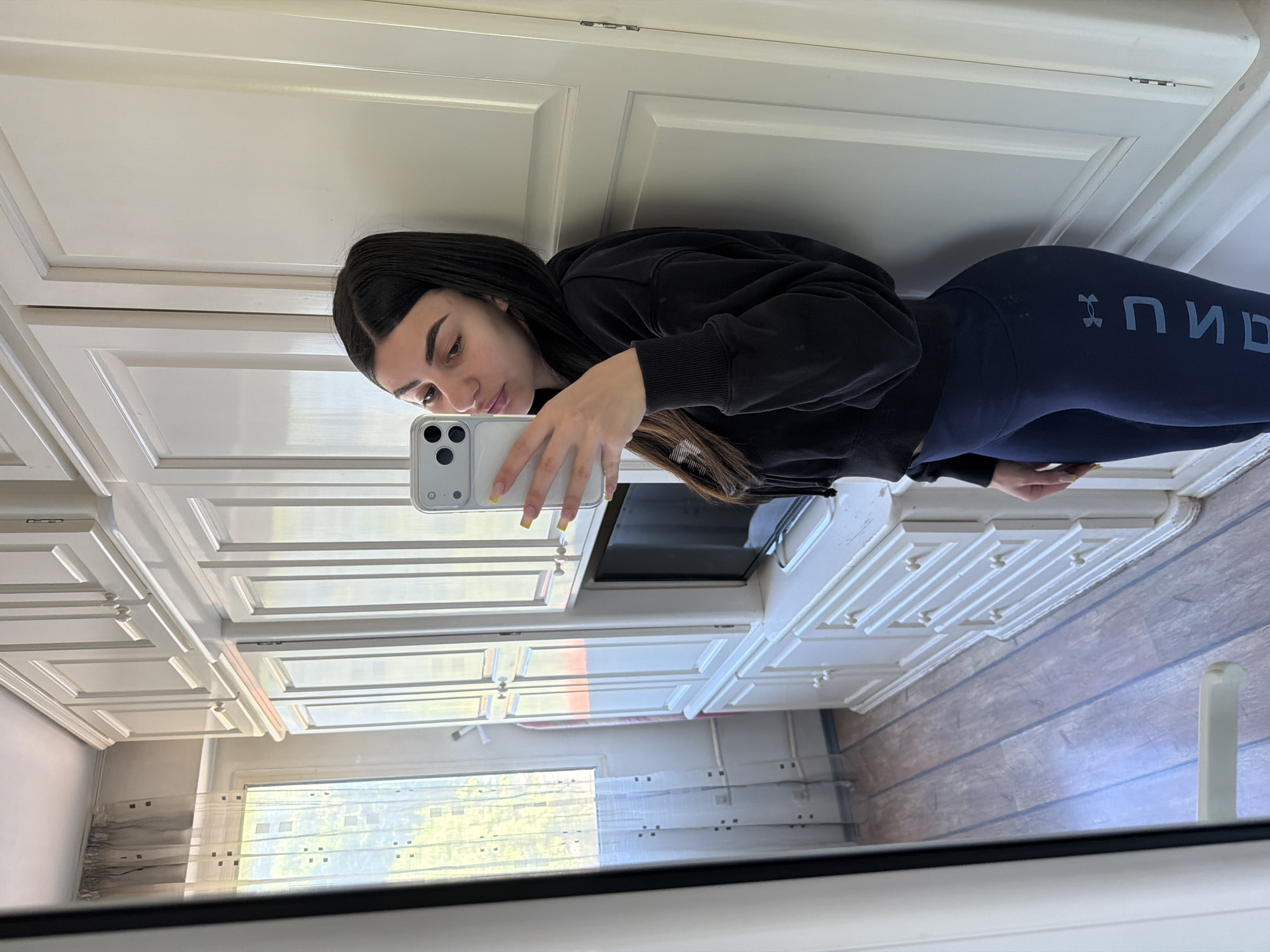 A young woman is taking a selfie in front of a mirror inside a room that appears to be a kitchen. She is wearing a black hooded sweatshirt and dark leggings with the word "UNDER ARMOUR" visible. She is holding a white smartphone. The background includes white kitchen cabinets, a window with a sheer curtain, and a glimpse of a building outside the window. The location cannot be determined from the information in the picture.
