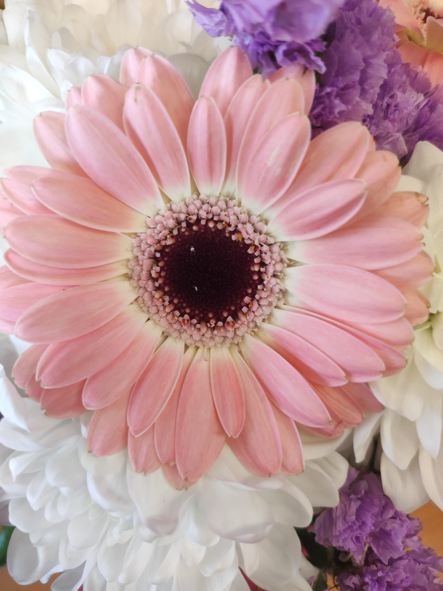The image shows a close-up of a flower arrangement. A large, light pink gerbera daisy dominates the center. Its petals are smooth and slightly curved. The flower's center is dark purple. Surrounding the gerbera daisy are smaller white and purple flowers with a feathery texture. The background is blurred, suggesting a shallow depth of field. There is no indication of this image being taken in the city of Votkinsk, Russia. No people, specific subjects, or actions are visible.