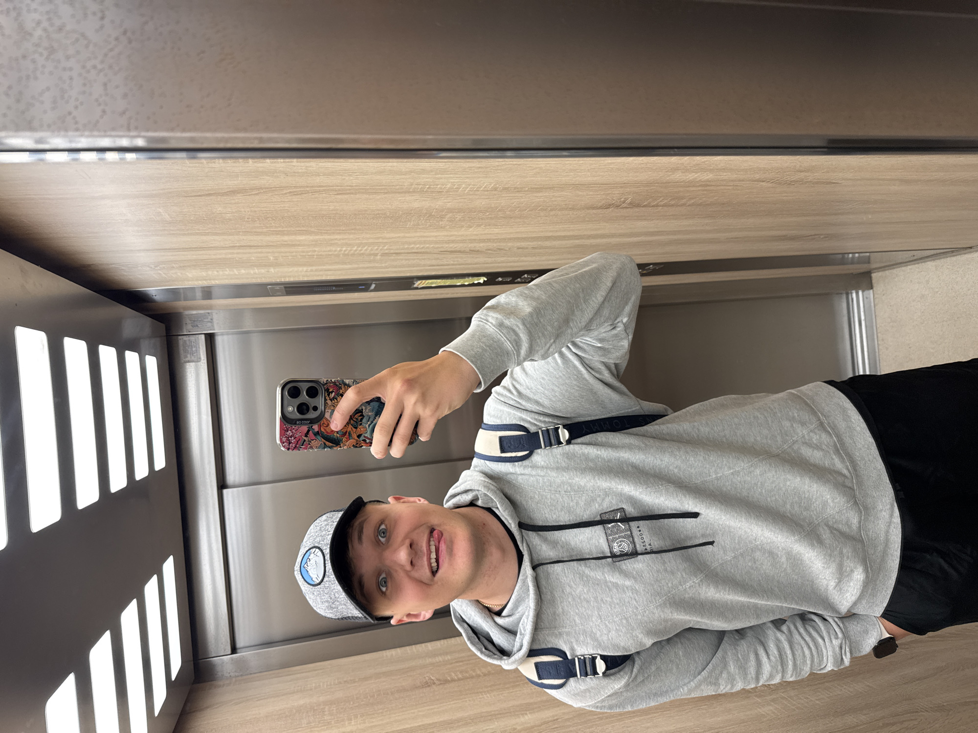 This image depicts a young man taking a mirror selfie inside an elevator, located in Plovdiv, Bulgaria. The man is captured from the chest up, positioned slightly off-center, and engages directly with the camera with a playful expression, his tongue extended slightly. He is dressed in a light grey hooded sweatshirt featuring black and cream-colored strap details, with the brand name "TOMMY" partially visible on the straps. He also wears a grey and white baseball cap adorned with a blue and white mountain-themed circular logo. In his right hand, he holds a smartphone encased in a vibrant, colorful, patterned case, on which the text "SO COOR" is partially discernible.
The elevator's interior showcases light wooden wall panels complemented by sleek metallic accents. To the left, a wall is fitted with multiple bright, rectangular illuminated panels, providing strong ambient lighting. The floor appears to be a light-colored wood or laminate material. The scene conveys a casual and lighthearted moment.