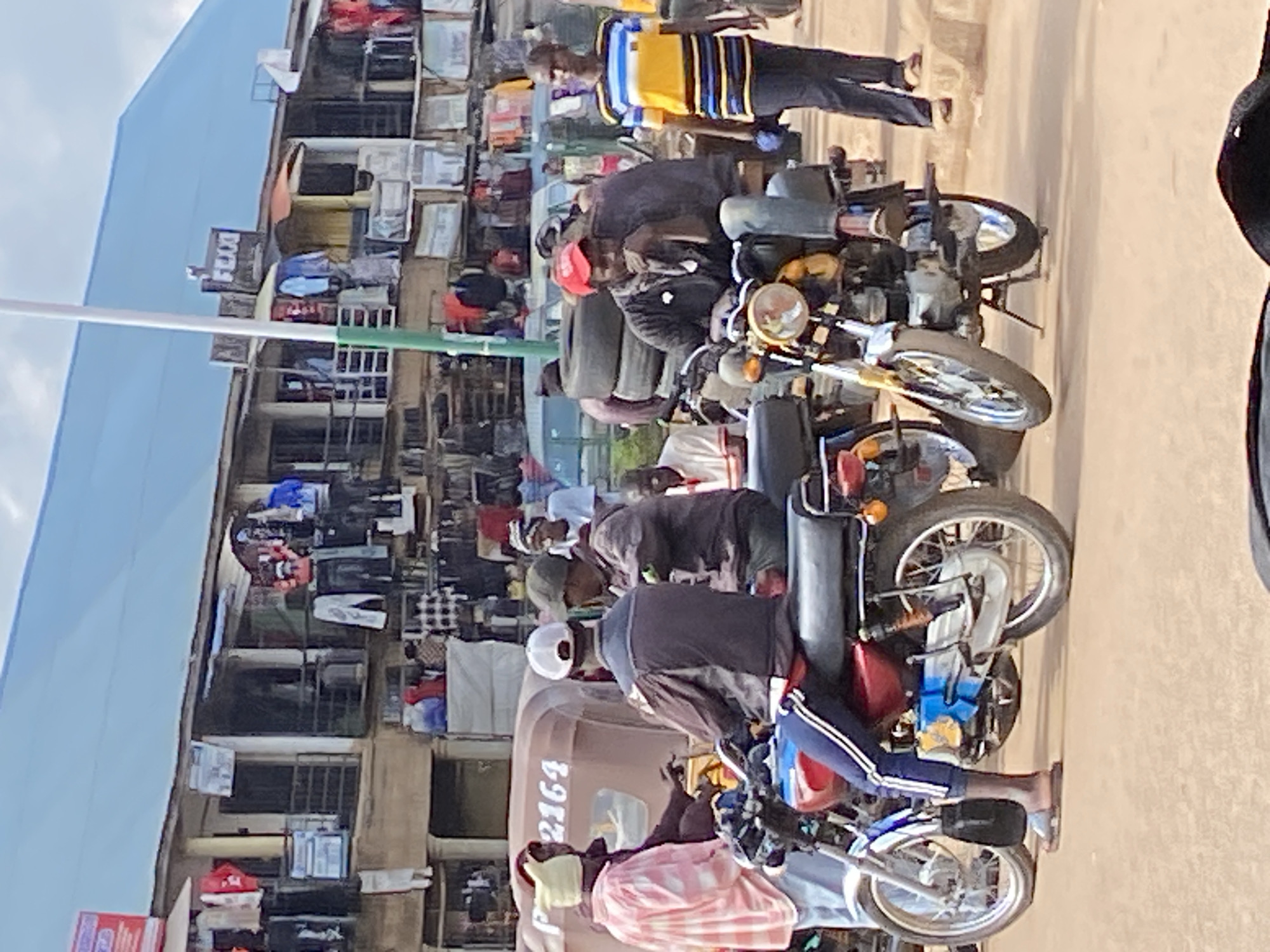 In the city of Bukuru, Nigeria, a street scene unfolds under a bright sky, likely mid-morning or afternoon, given the lighting. The foreground is dominated by two motorcycles. The motorcycle on the left is red and silver, with a rider whose lower body is visible, wearing blue pants with white stripes and brown sandals. He is seated on the motorcycle, which has a black seat and a blue and yellow detail on its side. A black bag hangs from the side of this motorcycle.
To the right, another motorcycle, predominantly yellow and silver, is parked. A man wearing a red cap, dark long-sleeved shirt, and blue jeans is seated on this motorcycle, resting his chin on his hand and looking down. He has a black lanyard around his neck. Behind him, a stack of tires is visible.
In the background, a three-wheeled vehicle, possibly a tuk-tuk, is partially visible with the license plate "P 2164" clearly displayed on its side. Several people are interacting around this vehicle. One individual in a pink and white checkered shirt and yellow head covering appears to be speaking to another person in a dark jacket. Further back, other individuals are standing and walking.
The environment suggests a marketplace or busy street with buildings lining the background. These buildings have multiple levels with balconies and are adorned with clothing items displayed for sale, hanging from railings and stalls. Signs with text, including "FLEXX," are visible on the structures, indicating commercial activity. The ground is a dirt or unpaved surface, common in many Nigerian urban settings. The overall scene depicts a typical day in a bustling Nigerian town, with people engaged in daily activities involving transportation, commerce, and social interaction.