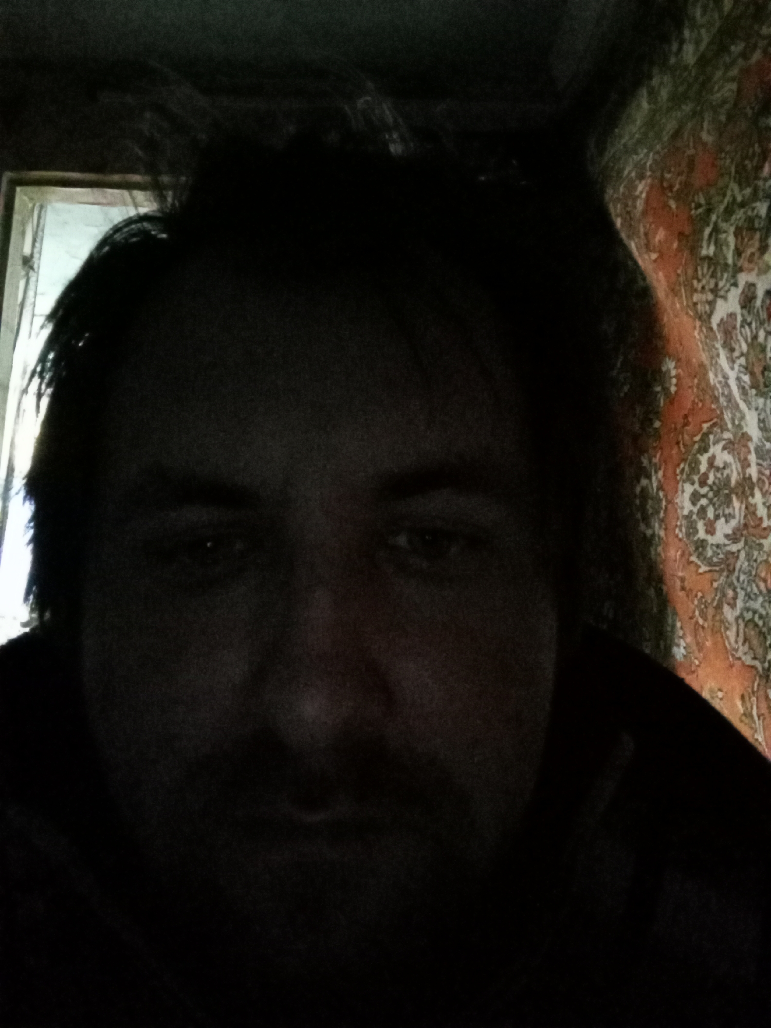 A dimly lit close-up photograph features a person with dark hair and facial hair. The background includes a partially visible patterned tapestry in shades of orange and green on the right side, and a blurred frame of a window or doorway on the left side. The location appears to be indoors, potentially in a private residence. No specific information is available to indicate the location is in Parabel, Russia.
