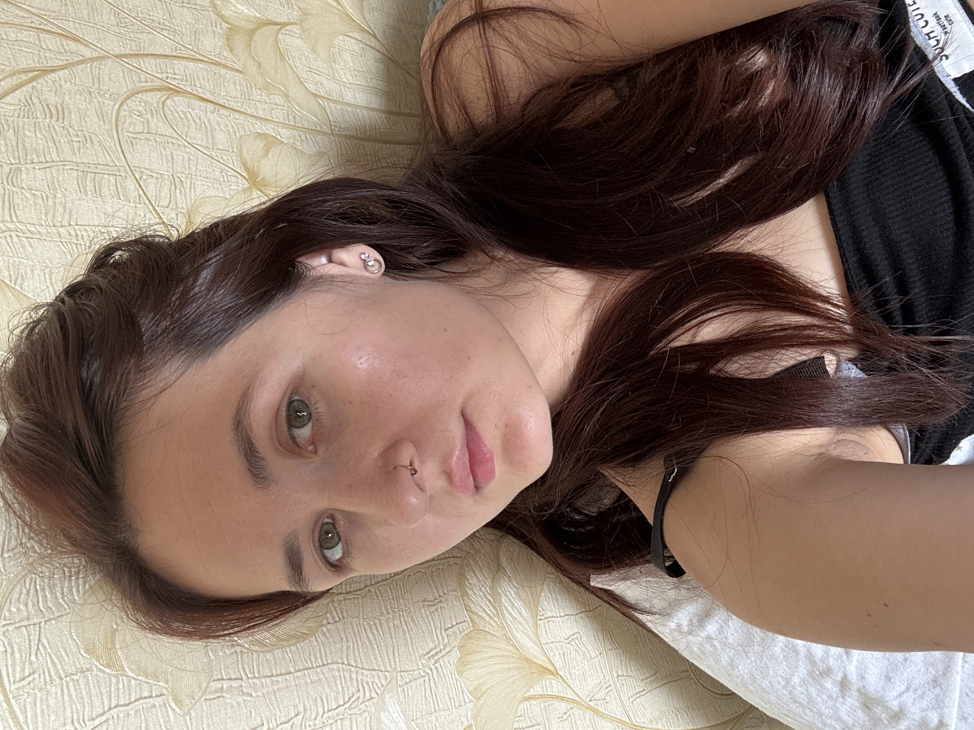 This is a close-up, high-angle selfie of a young woman lying down, likely in a bedroom in Naberezhnye Chelny, Russia. The woman has fair skin, green eyes, and long, dark brown hair. She is wearing a black top. Her left ear has a small, silver stud earring with two circles and a small pink stone. She also has a gold hoop nose ring. The background appears to be a wall with a textured floral wallpaper in a pale yellow or cream color. A white pillow is partially visible in the lower left corner. The lighting suggests it is daytime, possibly near a window, casting a soft glow on her face. The image is focused on the woman's face and upper body.