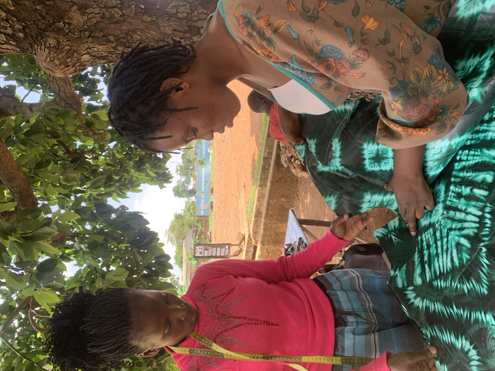 Two women are engaged in a tailoring activity outdoors in Maigizo, Nigeria. One woman is seated on a patterned cloth, wearing a tan, floral-patterned top with a white undershirt. The other woman is standing, wearing a pink sweater, striped bottoms, and a measuring tape around her neck. They are examining a piece of green tie-dye fabric. The scene is shaded by a tree. In the background, there is a dirt area, buildings, and a sign that says “POS agent.”