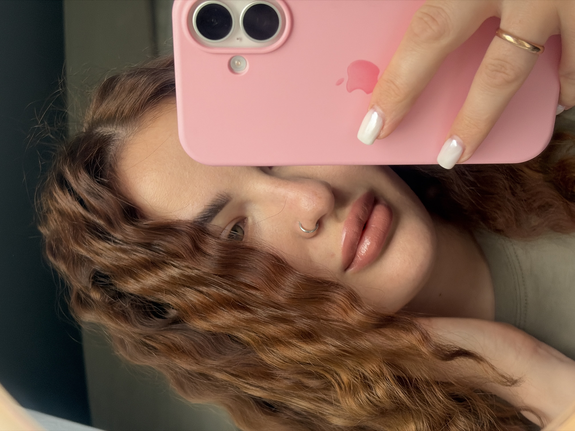 A woman with light skin and auburn, wavy hair is holding a pink iPhone up to her face, partially obscuring it. The phone has two cameras and an Apple logo on the back. The woman has light-colored nails, one of which rests on the phone near the logo, and wears a gold ring on another finger. A silver nose ring is visible. The background is blurred and indistinct, suggesting an indoor setting. No location in Novopokrovskaya, Russia can be determined from the image.
