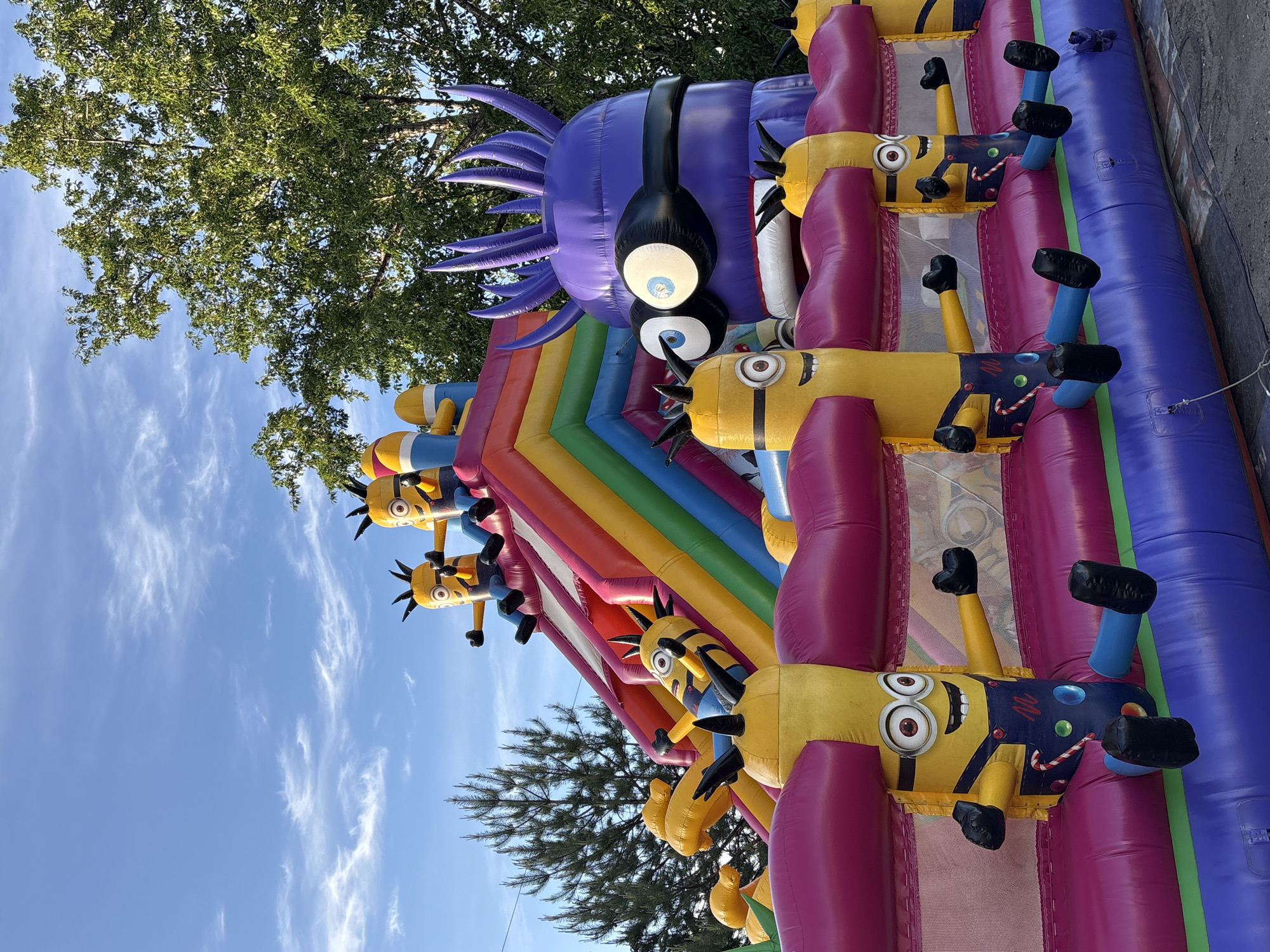The image shows an inflatable bounce house with a Minions and purple monster theme. The bounce house has a rainbow-colored slide and several inflatable Minion figures attached to its sides. The sky is blue with scattered clouds, and trees are visible in the background. The image does not contain any information about the location, city of Birobidzhan, Russia, people, subjects, objects, actions, or interactions.