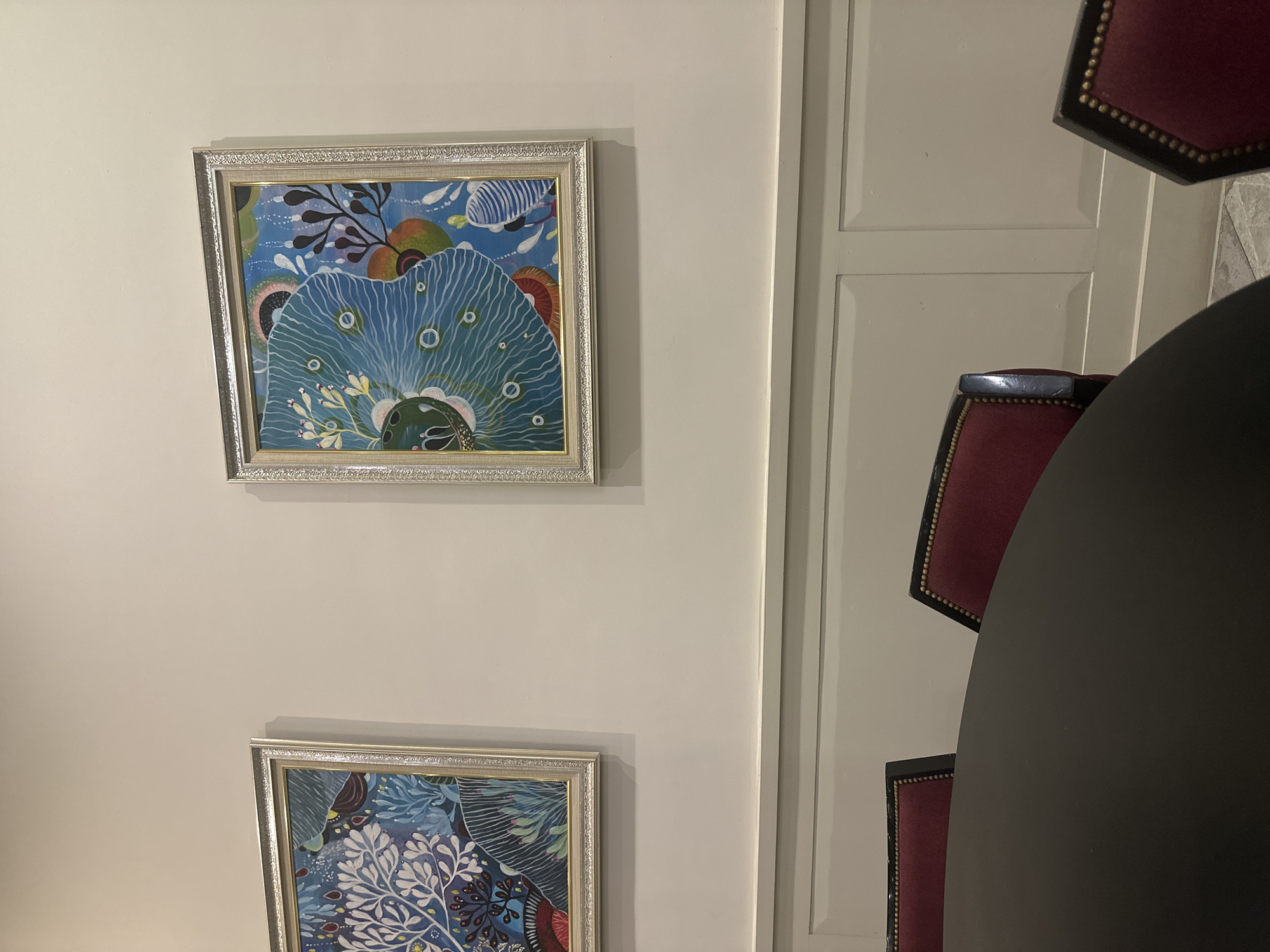 The image depicts an indoor scene with a cream-colored wall featuring two framed paintings with silver frames. The paintings appear to have floral or organic designs in blue, white, and other colors. To the right of the wall is a section of a door or wall panel painted in a similar cream color with vertical recessed panels. A black round table is visible, partially obscured by the right side of the frame. Around the table, there are red chairs with dark wood trim and studded detailing. The location is unknown.