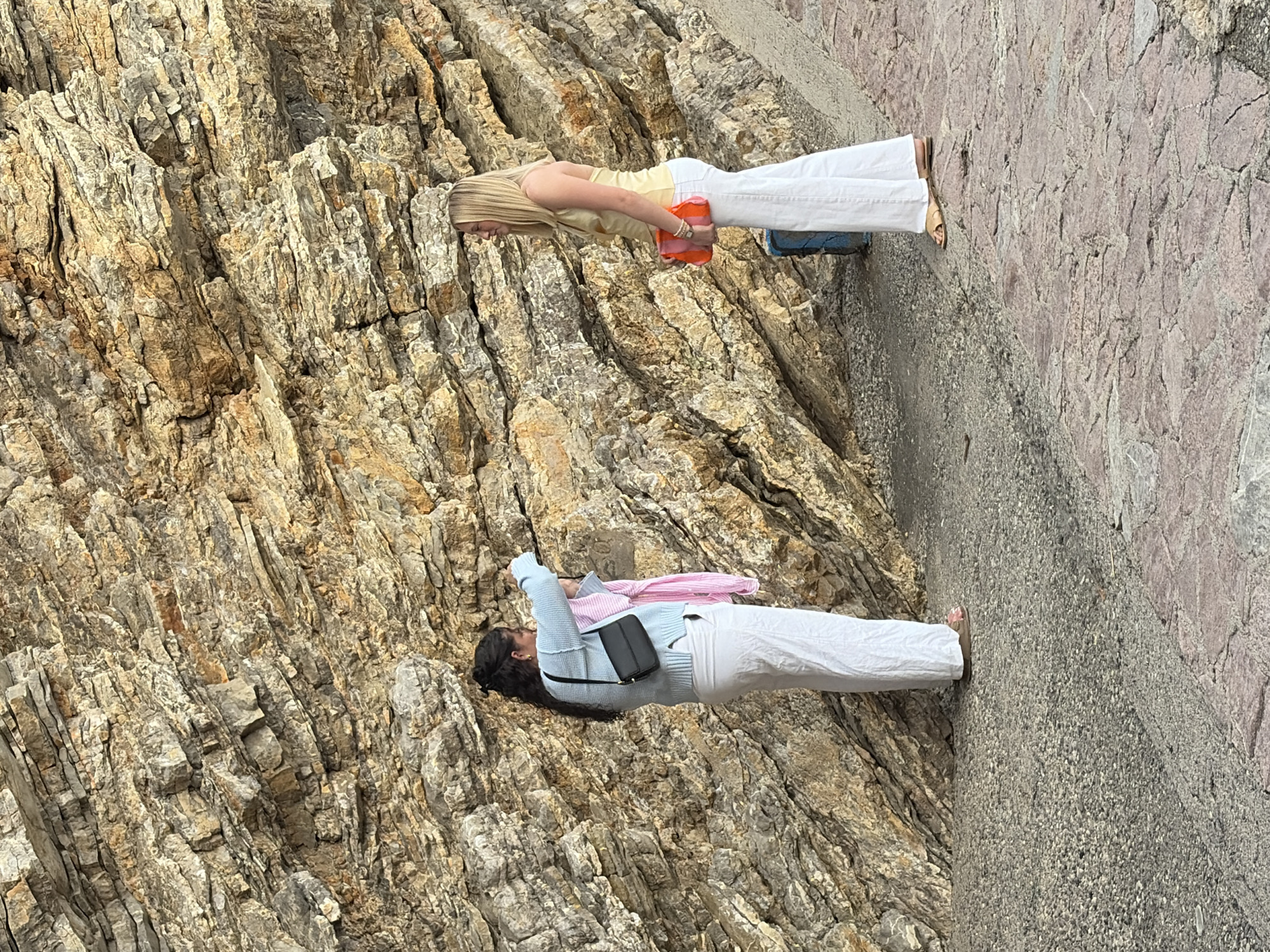 Two women are standing on a textured stone surface, likely a walkway or observation point, adjacent to a large, steeply angled rock face. The rock face, taking up the left side of the frame, has a complex, layered appearance with various shades of brown, gray, and beige.
One woman has blonde hair, wears a yellow top and white pants, and is holding a pink object. The other woman has dark hair pulled back in a bun, wears a light blue top, white pants, a black purse, and has a pink garment draped over her shoulder.
The background and stone textures suggest this location is in a natural or semi-natural environment, possibly near a coastal area. Given the context of Budva, Montenegro, the scene is likely located at a site near the rocky cliffs typical of the Budva Riviera.