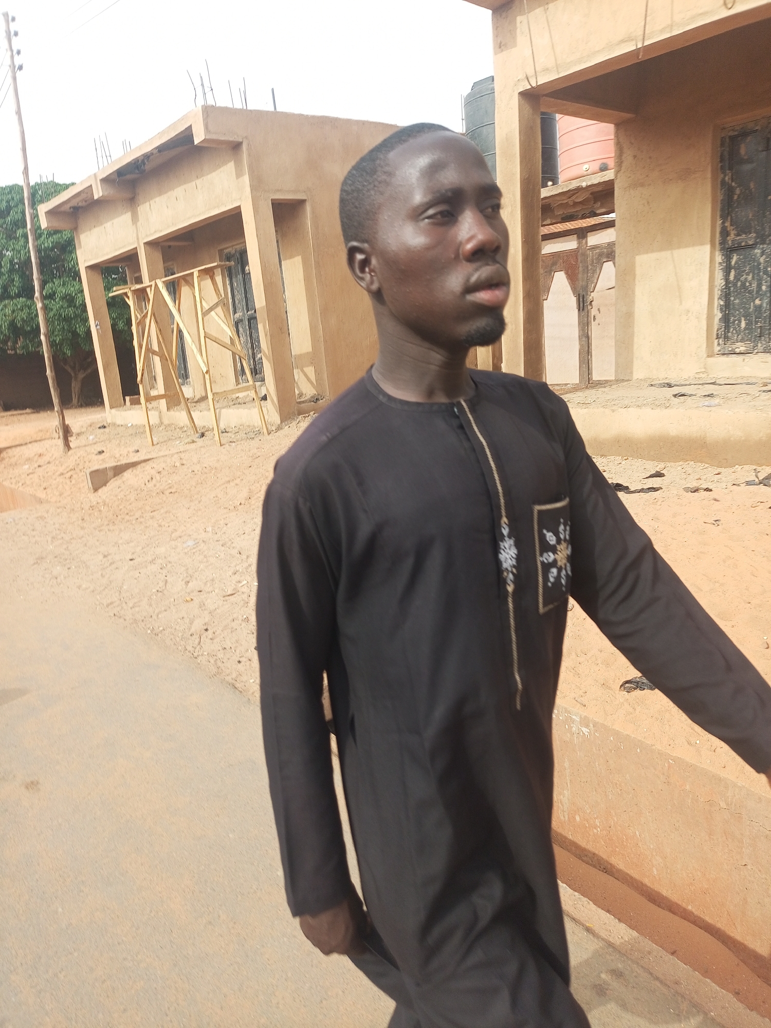 A man is standing on a paved surface near what appears to be a residential area in Gombe, Nigeria. He is wearing a long-sleeved, dark-colored garment with decorative detailing on the chest. Behind him are buildings constructed of tan or light brown materials. One building seems to be under construction, indicated by wooden scaffolding. The ground has sandy areas and the sky is overcast. Water tanks are visible on the roofs of some of the buildings.