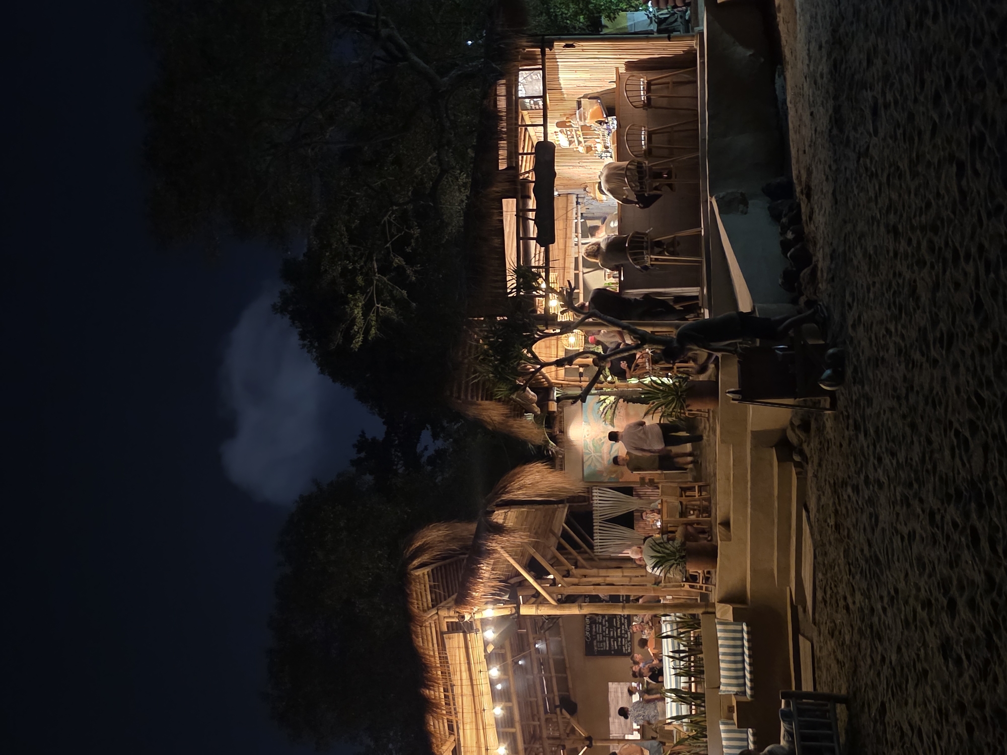 A night shot shows a multi-level, open-air restaurant situated on a beach. The restaurant structure is primarily made of bamboo and thatch roofing. At the top level, bar stools are visible, indicating a bar area. Lower levels feature tables and chairs occupied by patrons, suggesting a dining area. The atmosphere is illuminated by warm, artificial lighting. The beach, a sandy expanse, is adjacent to the restaurant. Dark trees are visible near the restaurant. A faint patch of light, potentially a moon, can be seen through the trees. The city of North Kuta, Indonesia is not directly visible but assumed from the geographic context.