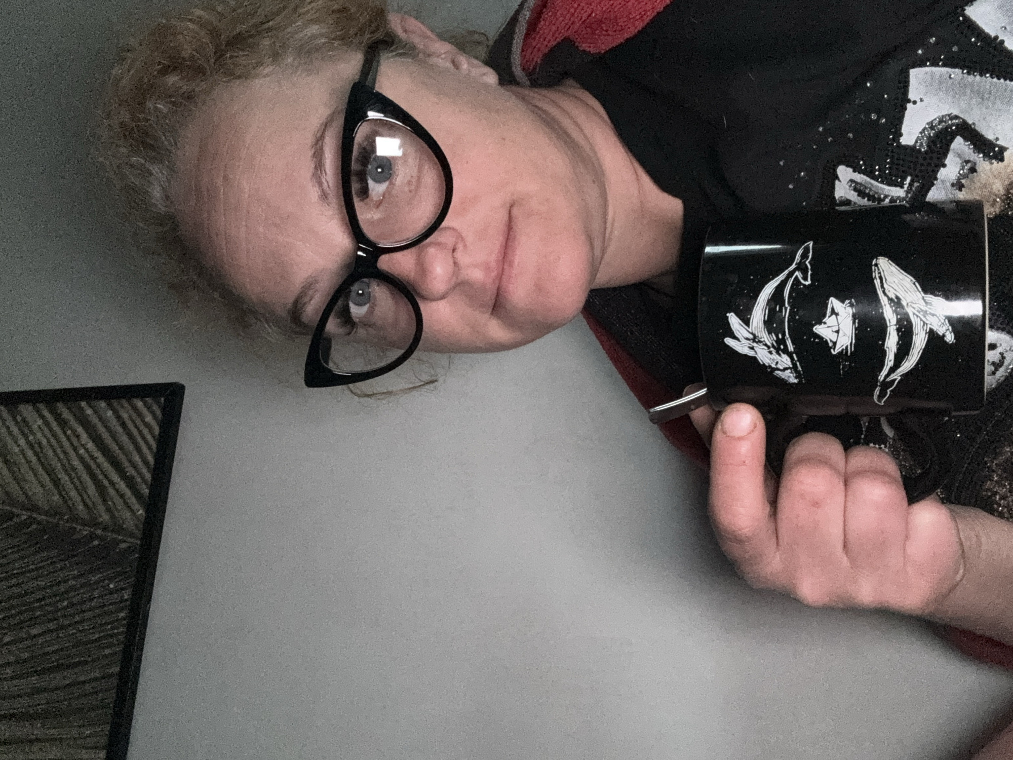 A selfie-style, low-angle shot features a woman with fair skin and blonde hair wearing black-rimmed glasses. She holds a black mug with a white design featuring two whales and a paper boat, her right hand gripping the mug handle. The background includes a gray wall and part of a dark-framed picture. The location is not specified in the image and cannot be determined.
