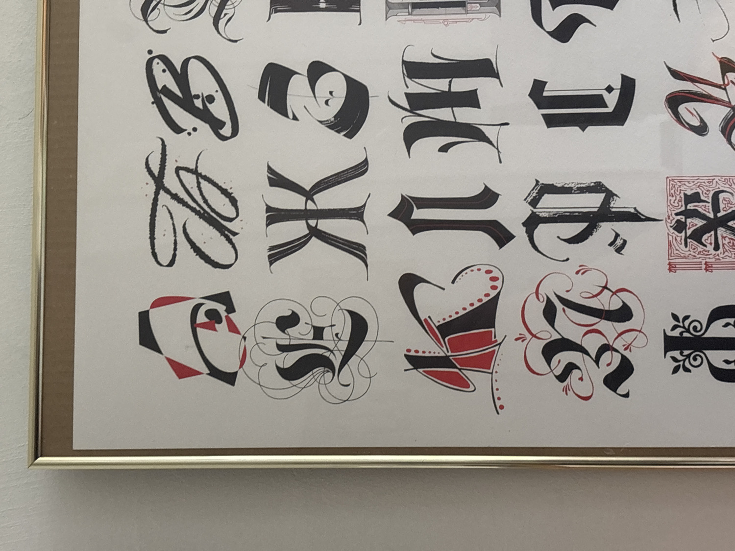 A framed print featuring an extensive collection of calligraphic capital letterforms hangs on a plain, light-colored wall. The frame is slender and appears to be made of a metallic gold or brass material. The print itself showcases various historical and decorative typographic styles, predominantly in black ink with significant red accents and outlining.
The artwork is arranged in multiple rows of large, stylized letters and ornate designs. Visible examples include:
- A gestural 'B' or similar character with expressive brushstrokes and speckled details.
- A flowing, elegant 'L' crafted with sweeping calligraphic lines.
- A bold, angular 'K' with broad, clean strokes.
- Several letters in a Gothic or Blackletter style, such as an 'H' and an elongated 'I', characterized by heavy downstrokes and sharp serifs.
- A complex, almost block-like character with textured strokes, possibly an 'E' or 'F'.
- An abstract 'C' composed of geometric black and red shapes.
- An ornate 'S' with elaborate red and black flourishes and swirls, indicative of decorative script.
- A distinctive character (possibly 'M' or 'N') with a heart-like shape, adorned with red dots.
- A highly decorative 'R' or 'B' featuring heavy black strokes intertwined with intricate red swirls.
- A heavily embellished 'P' in a Gothic-revival style, incorporating detailed floral patterns.
The diverse array of letter styles, from traditional Gothic to modern geometric and fluid calligraphic, demonstrates a wide range of artistic interpretations. No specific text or words are legible, as each letter is presented as an individual art piece. The overall impression is one of artistic typography and design.