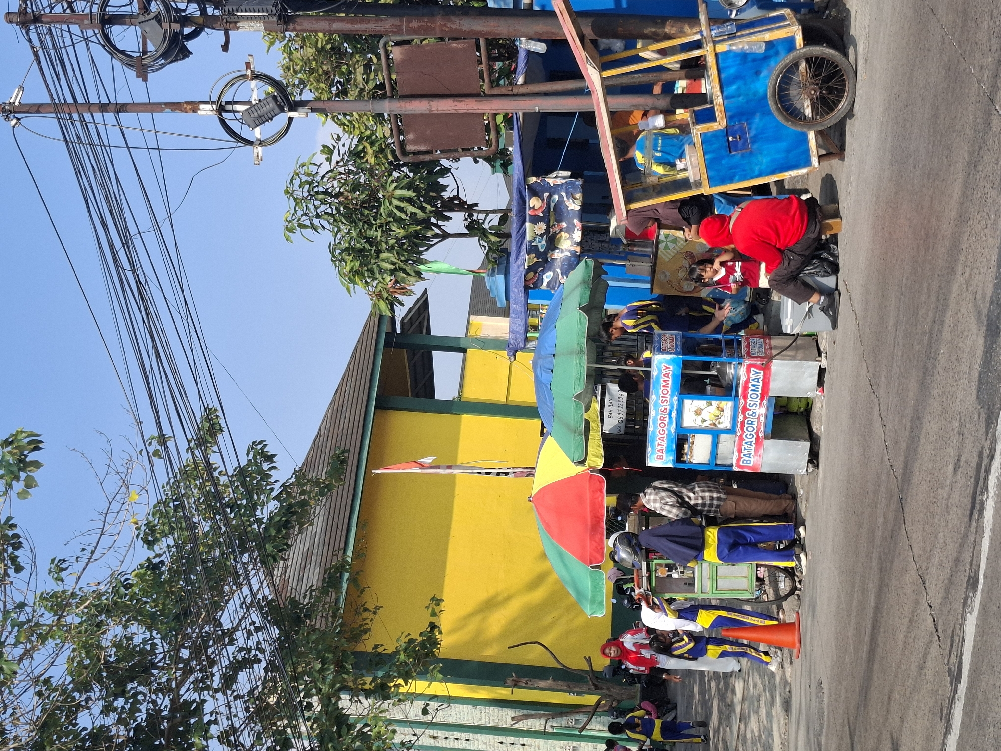 The image depicts a street scene in Sukabumi, Indonesia, featuring several food carts and a building. In the foreground, a blue and yellow food cart with a large wheel is parked. To the left of this cart, there are two other food carts, one with a red and green umbrella and another with a green and blue umbrella. These carts are selling food, with signs indicating "BATAGOR & SIOMAY". Several people are visible around the food carts. A person in a red hooded sweatshirt is sitting on a stool near the blue and yellow cart, facing away from the camera. Other individuals, some wearing school uniforms (blue and yellow), are standing and interacting with the vendors. Behind the food carts, a brightly painted yellow building with green trim is visible, and a red and white Indonesian flag is flying from a pole. The street is paved, and there are overhead power lines and a utility pole on the right side of the image. The sky is clear and blue.