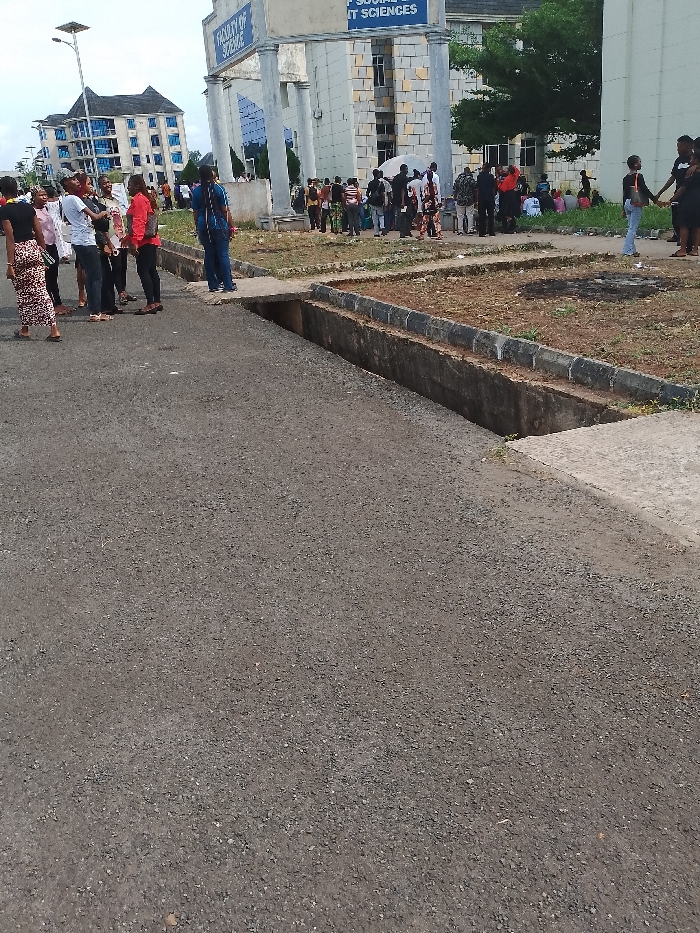 The scene depicts an exterior view in Umueshi, Nigeria, likely on a university campus. In the background, a building is labeled "Faculty of Social and IT Sciences" and "Faculty of Science." A group of people are gathered near the building. In the foreground, there's a paved area with a concrete drainage channel. The ground is dry and appears worn. The day appears overcast with diffused light.