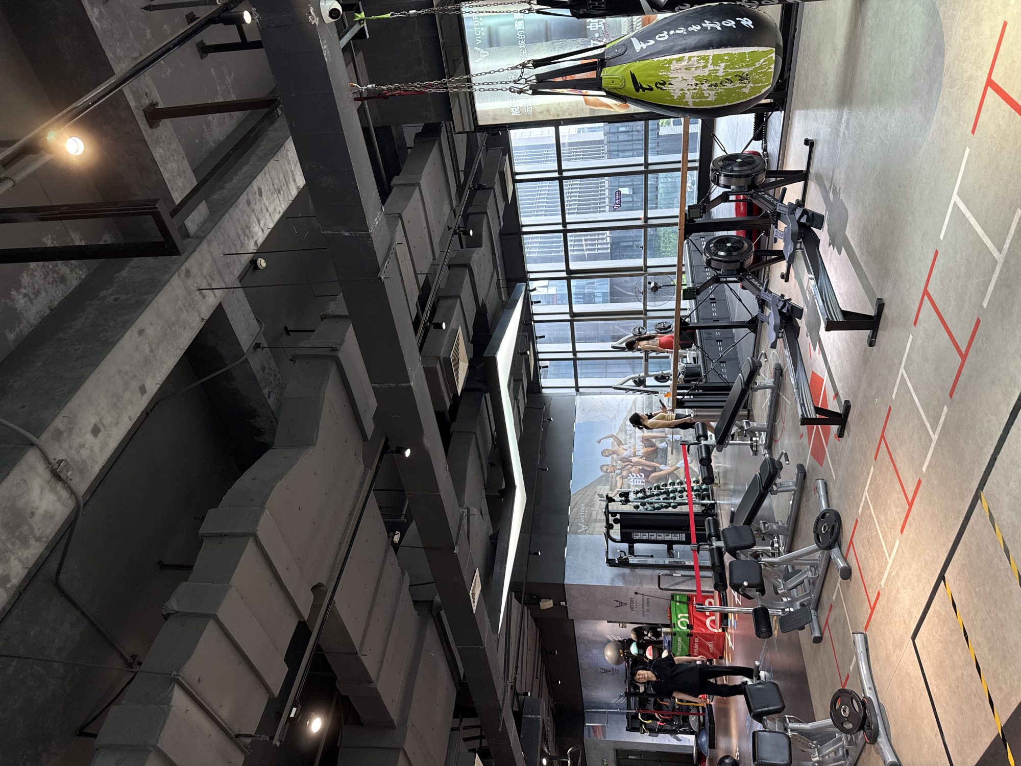 The image shows the interior of a modern gym located in Haizhu District, China. The gym features an industrial-style ceiling with exposed concrete and ductwork, with various lights and a security camera visible. The floor is marked with lines in red, white, and yellow, indicating different workout zones.
In the foreground, several pieces of exercise equipment are present, including rowing machines, weight benches, and weight plates on the floor. A large green and black punching bag hangs from the ceiling on the right side of the frame.
In the midground, two women are visible. One woman, wearing black athletic wear, walks towards the left side of the image. Another woman, in a yellow tank top and red leggings, stands near a weight rack on the right. Several other people are partially visible in the background.
The gym is equipped with various weight machines, exercise balls, and other fitness equipment. A large poster with images of athletes and text in Chinese adorns a wall in the background. The gym has large windows that offer a view of city buildings.