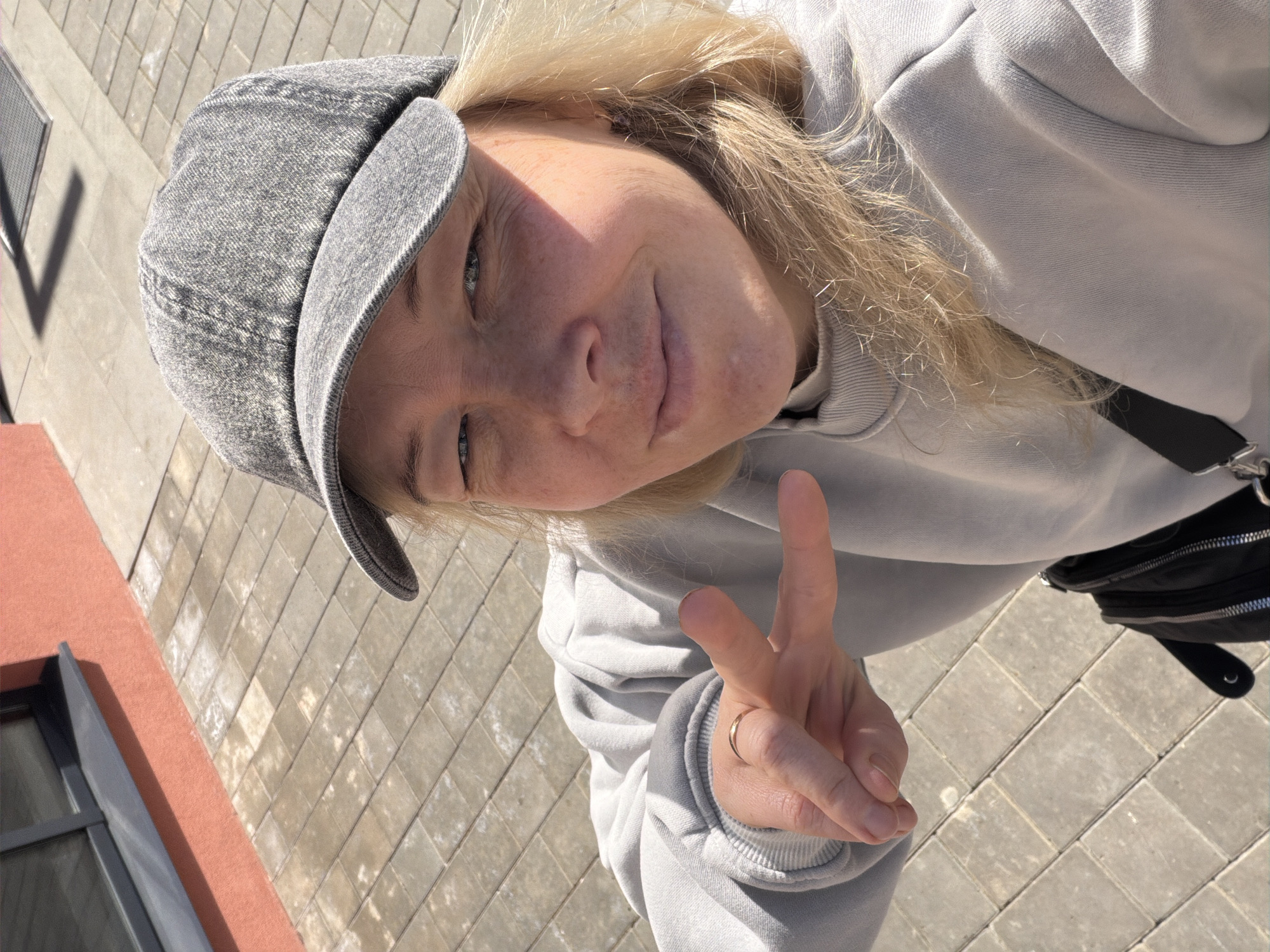 A blonde-haired woman wearing a gray cap and a gray hoodie is seen making a peace sign with her right hand. The woman is also wearing a gold ring. The background includes a brick-paved area and a building with a visible window frame, painted red. The image is rotated. The location is possibly Pervomaysky, Russia.