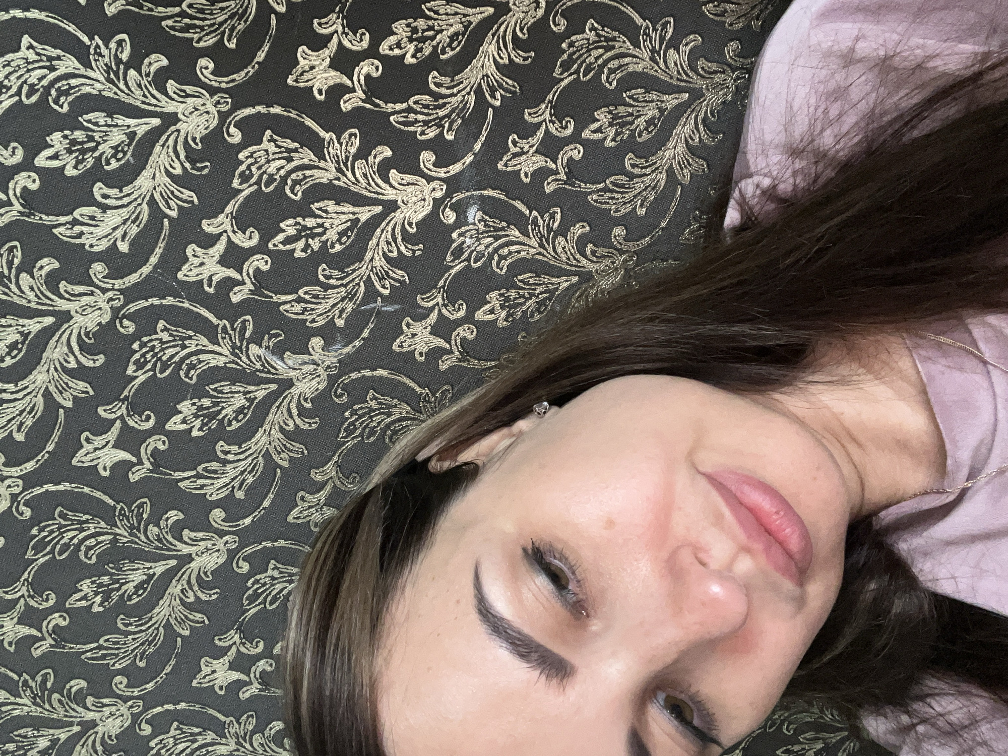 A medium close-up selfie of a woman with long dark brown hair, who is looking down and to her left. She has fair skin, dark eyebrows, and is wearing a light pink shirt and a silver necklace. Her face is slightly flushed, and she is wearing light makeup, including eyeliner and lipstick. She has a small silver stud earring in her visible ear. The background is a dark wallpaper with a gold damask pattern. The image is cropped at the woman's right shoulder and the top of her head.