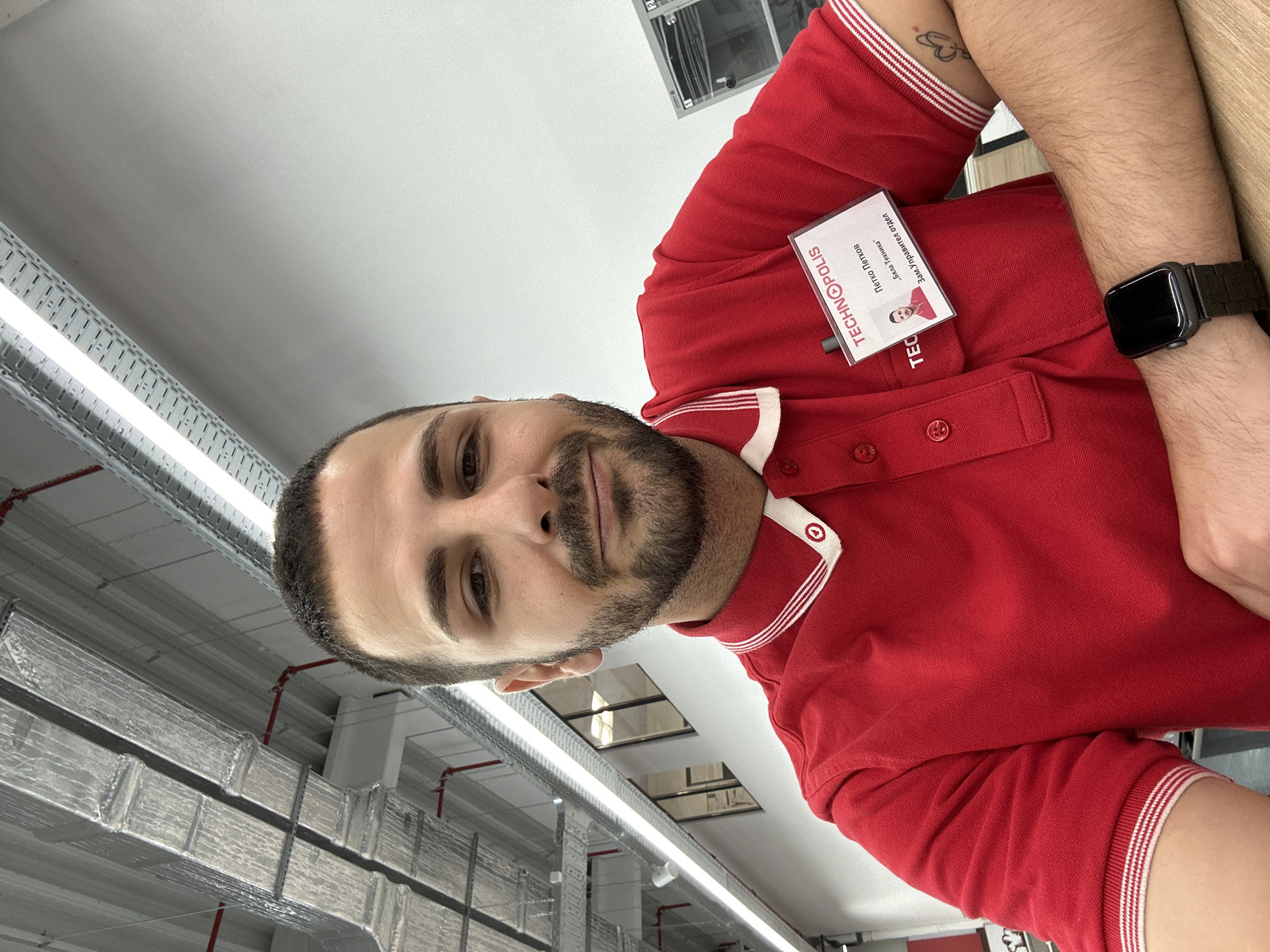 A man is shown from the chest up, wearing a red polo shirt with white trim around the collar and sleeves. He has a name tag that says "TECHNOPOLIS" and features a photo of him. The man has dark hair, a beard, and is wearing a smart watch on his left wrist. In the background, there are industrial-looking ceilings with metal beams, pipes, and overhead lighting.
