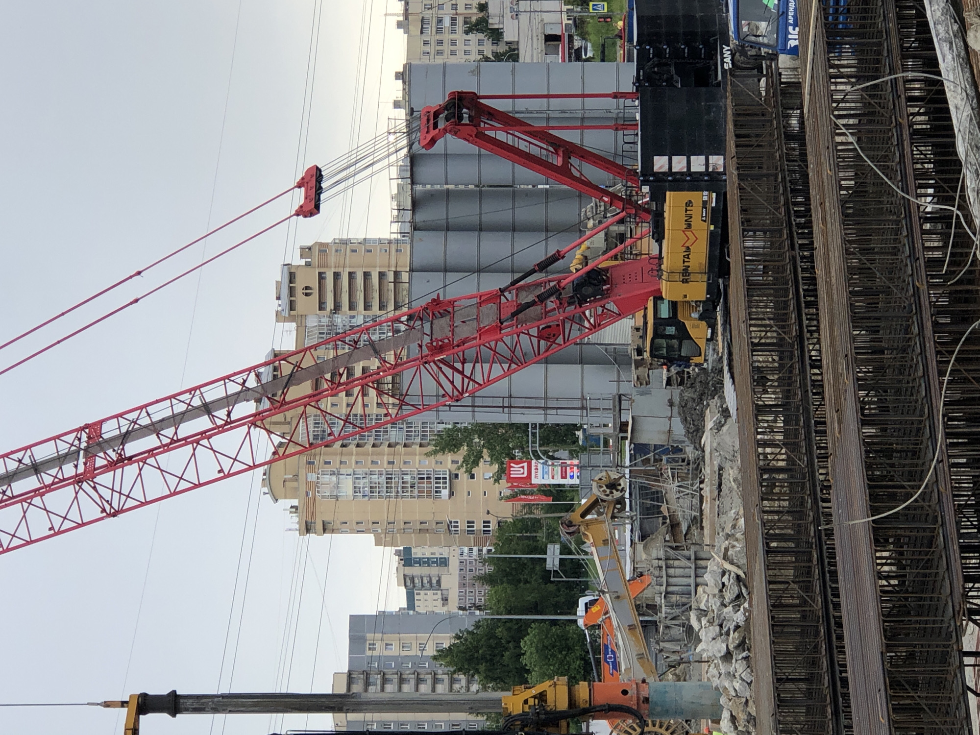 An overcast sky looms over a construction site in St. Petersburg, Russia. A massive red crane dominates the frame, its boom extending diagonally upwards. To the left, a tall, grey cylindrical drilling rig stands against the sky. In the middle ground, several large, grey cylindrical silos are visible, with a yellow excavator positioned in front of them. The excavator has "RENTAL UNITS" written on its side. In the foreground, layers of rebar, forming cages for concrete reinforcement, are stacked horizontally. Behind the rebar, a pile of rubble and construction debris is present. The background features multi-story residential buildings, suggesting an urban setting. A red sign with a white logo and Cyrillic text, possibly advertising a company, is also visible. Power lines crisscross the sky above the construction site.