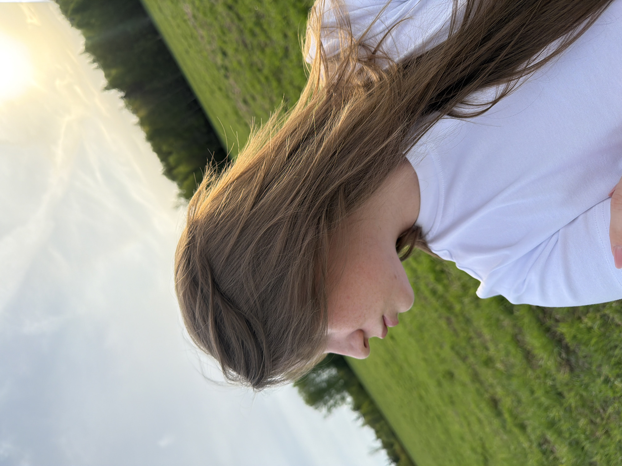 A young Caucasian girl with light brown hair and fair skin stands in a grassy field under a partly cloudy sky. She wears a white t-shirt. In the background, there are trees. Location is unknown, so can't relate it to Zlodar', Russia.