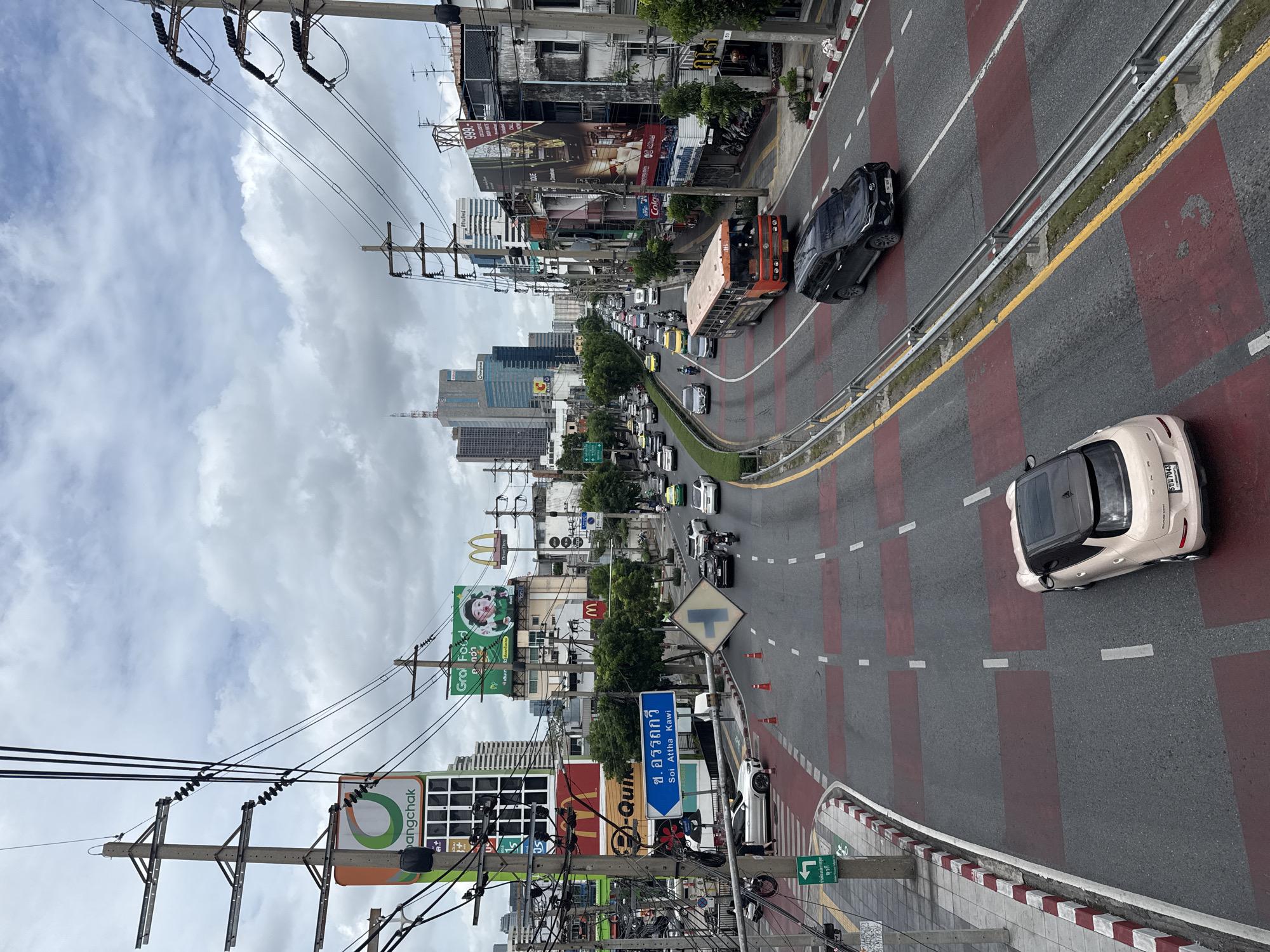 The image depicts a busy street scene in Bangkok, Thailand, likely during the daytime under an overcast sky. The perspective is from an elevated viewpoint, looking down at a multi-lane road filled with various vehicles. In the foreground, a light-colored car is parked, and further down the road, a dark SUV is in motion. An orange bus is also visible, as are numerous other cars, motorcycles, and a green taxi.
Dominating the cityscape are numerous power lines and utility poles, a common sight in Bangkok. Billboards and signs are prominently displayed, advertising brands like Bangchak, Grab Food, and McDonald's. The architecture is a mix of modern high-rise buildings in the distance and older, lower-rise structures closer to the street, suggesting an urban environment that is a blend of development. The road itself has distinct red and gray patterned markings, possibly indicating a pedestrian crossing or a specific traffic management system. Pedestrians are visible on the sidewalk and crossing the street. Traffic appears to be moderate to heavy, with vehicles moving in multiple directions. The overall impression is one of a bustling, active city environment.