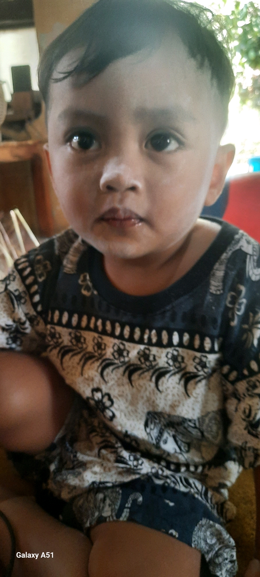 A young child, likely a boy due to the short haircut, is the central focus of this medium-shot photograph. He is looking directly at the camera with dark, round eyes and a slightly parted mouth. His skin appears fair, with some subtle redness on his cheeks and a dusting of white powder or residue on his forehead and nose. He has dark, short hair, styled with a slight wave on the left side.
The child is wearing a dark blue t-shirt with a patterned design. The pattern consists of a band of oval shapes and a repeated motif of stylized flowers with dark stems and leaves on a lighter, possibly off-white or beige background. The lower part of the shirt reveals a dark blue section with more intricate patterns, including what appear to be animalistic or human faces.
The background is blurred, suggesting an indoor or shaded outdoor setting. To the left, there is a glimpse of what might be wooden furniture and a light-colored object with vertical lines, possibly part of an appliance or structure. In the upper right corner, out-of-focus foliage is visible, indicating the presence of plants or trees. The lighting is somewhat diffuse, casting soft shadows.
The bottom left corner of the image displays the text "Galaxy A51," indicating the device used for capture. The child's legs and arms are visible, with his left arm resting on his lap and his right arm out of frame. The scene appears to be a casual, candid shot. The location is Ploso, Indonesia.