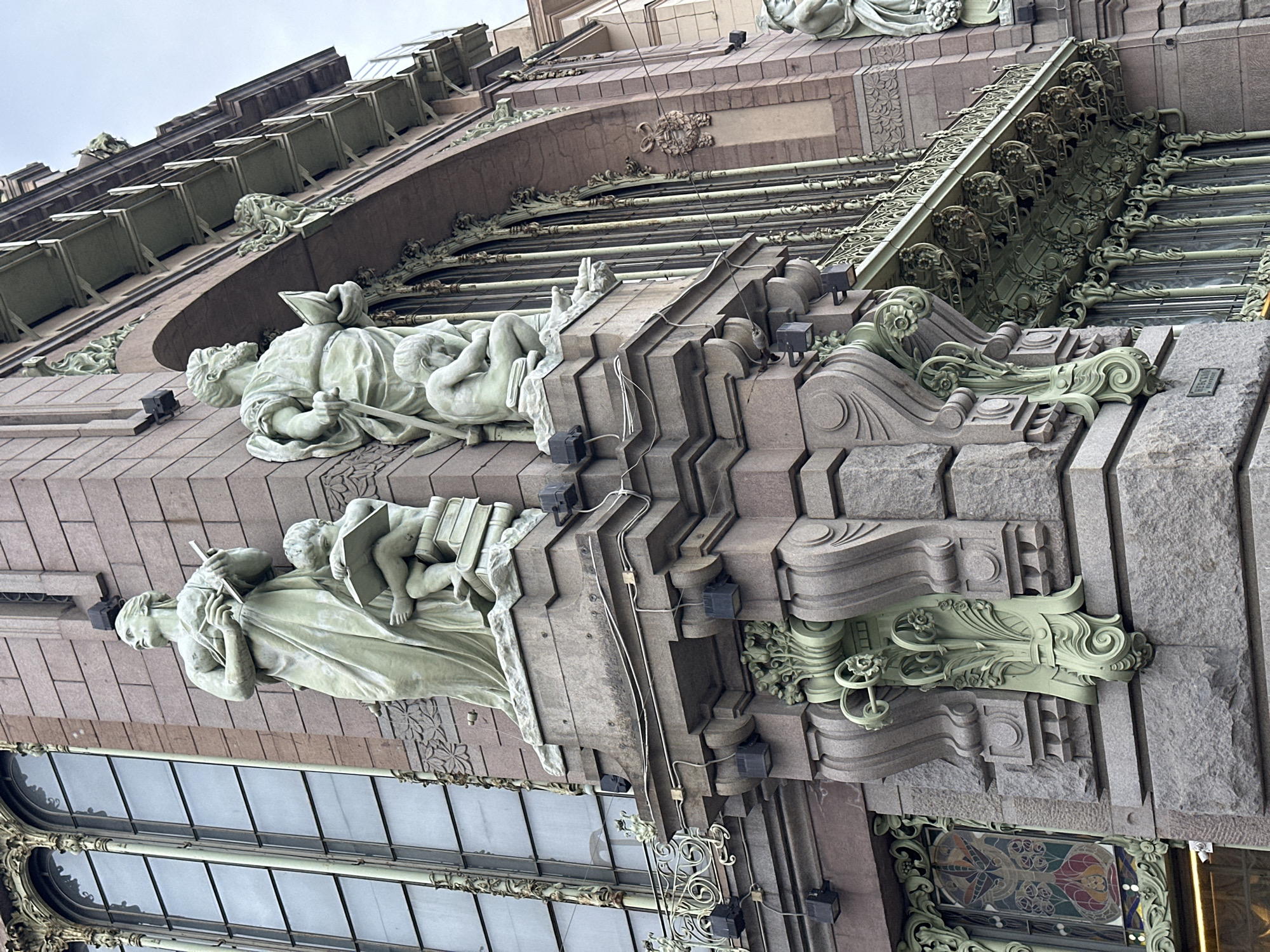 The image shows a section of the facade of the Eliseyev Emporium building in Saint Petersburg, Russia, specifically the Nevsky Prospekt side. Two pale stone statues of figures holding books are prominent. The building's exterior features intricate architectural details, including ornate window frames and decorative elements. The facade is composed of blocks of light brown and grey stone. There are visible electrical fixtures attached to the building, as well as an additional sculpture above the statues. The sky is overcast.