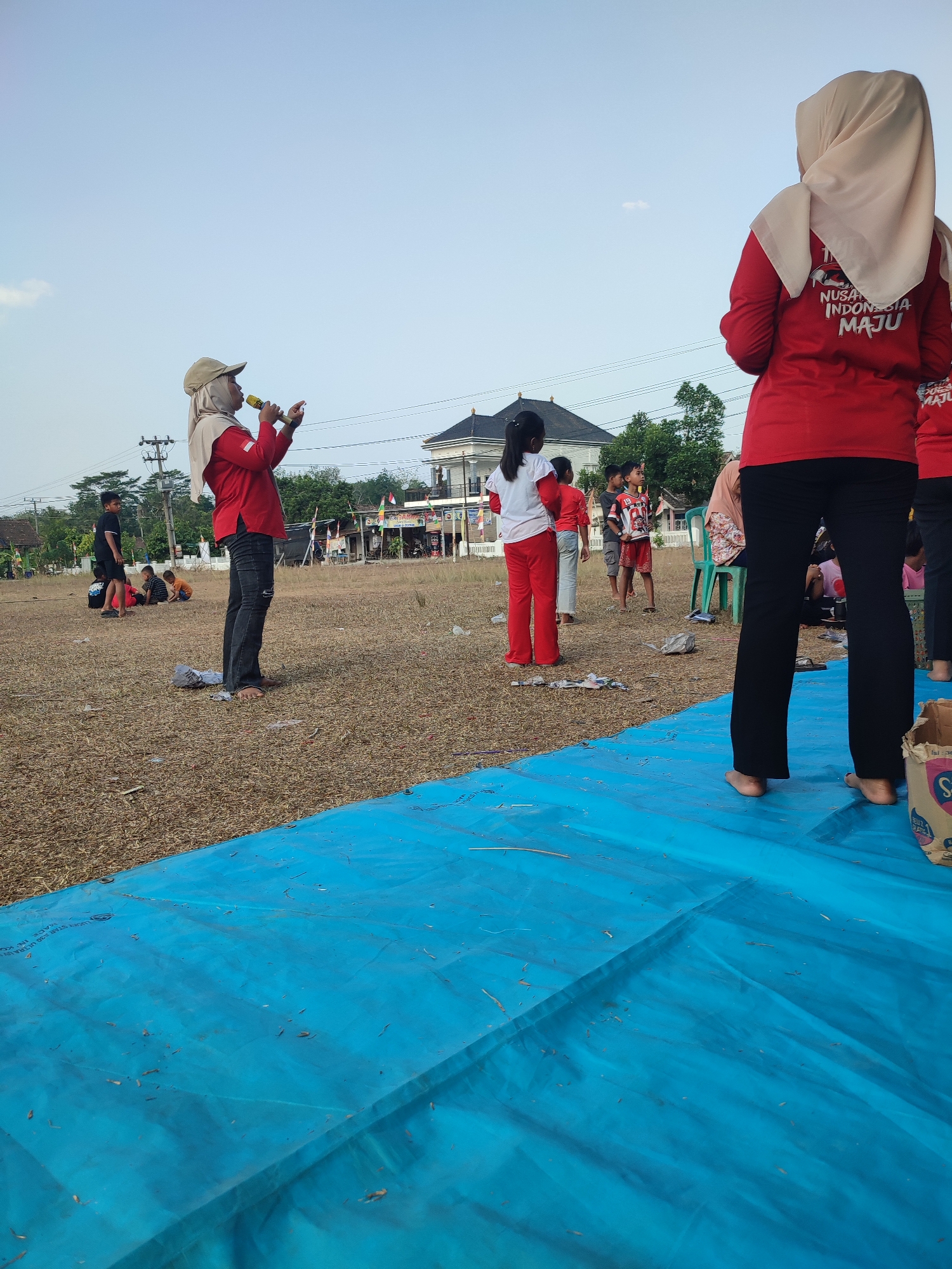 In Nusa Jaya, Indonesia, this outdoor scene captures a gathering on a dry, grassy field. A woman wearing a red long-sleeved shirt, black jeans, and a beige headscarf stands in the foreground, holding a microphone and speaking. She wears a beige baseball cap over her headscarf. Behind her, several people, including children, are visible. Some children are seated on the ground, while others stand and observe. One person, also in a red shirt with "NUSANTARA INDONESIA MAJU" printed on the back, is facing away from the camera. A large blue tarp covers part of the foreground. The background features trees, houses, and utility poles under a clear sky.