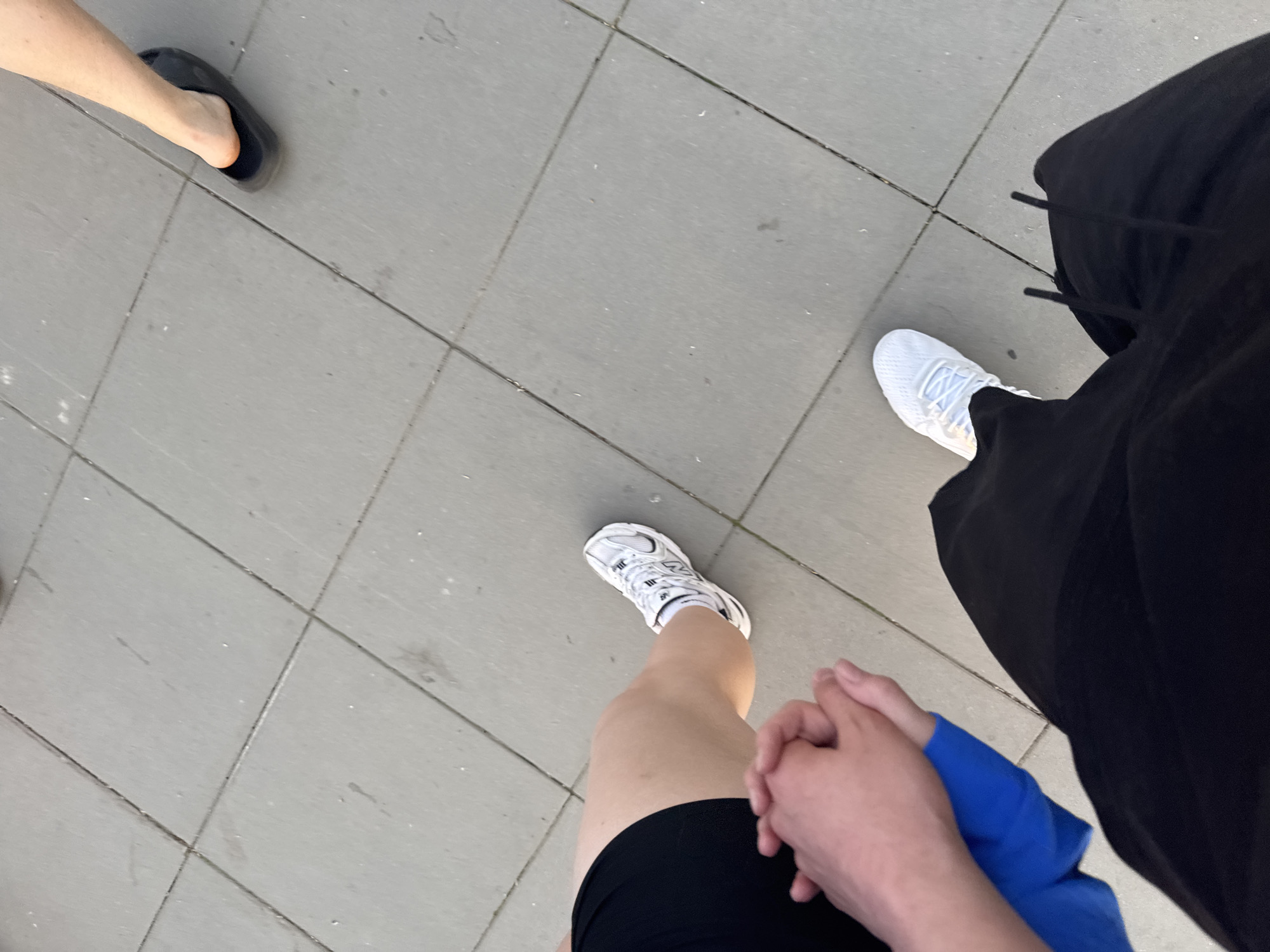 The image captures a street scene from an overhead perspective. The surface consists of square paving stones. Three individuals are partially visible. The person on the left is wearing a dark-colored sandal. The person in the center is wearing white athletic shoes with black shorts and appears to be holding hands with the person on the right. The individual on the right is wearing white athletic shoes and black sweatpants. The location is possibly Perm, Russia. The action involves walking and hand-holding. The environment is an urban setting with street paving.