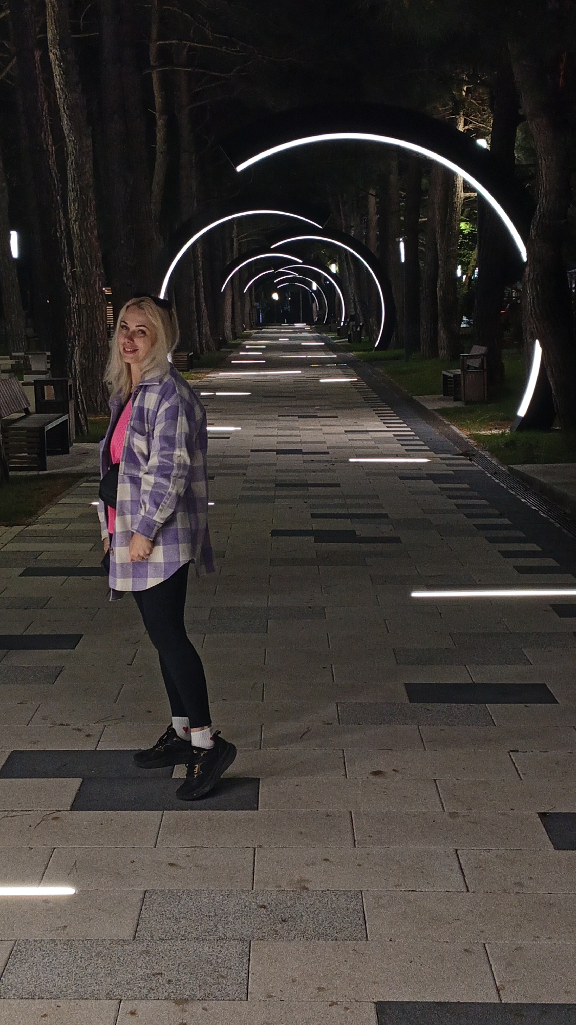A woman stands on a brick-paved walkway in Kabardinka, Russia. The walkway extends into the distance, lined with tall trees and illuminated by a series of glowing, semicircular light fixtures above, and horizontal light bars set into the pavement. The woman is facing the camera, dressed in black leggings, black shoes with white socks bearing small red hearts, a bright pink top, and a large, lavender-and-white checkered flannel shirt. She has a black shoulder bag. The environment includes benches along the walkway and additional vertical light fixtures near the trees.