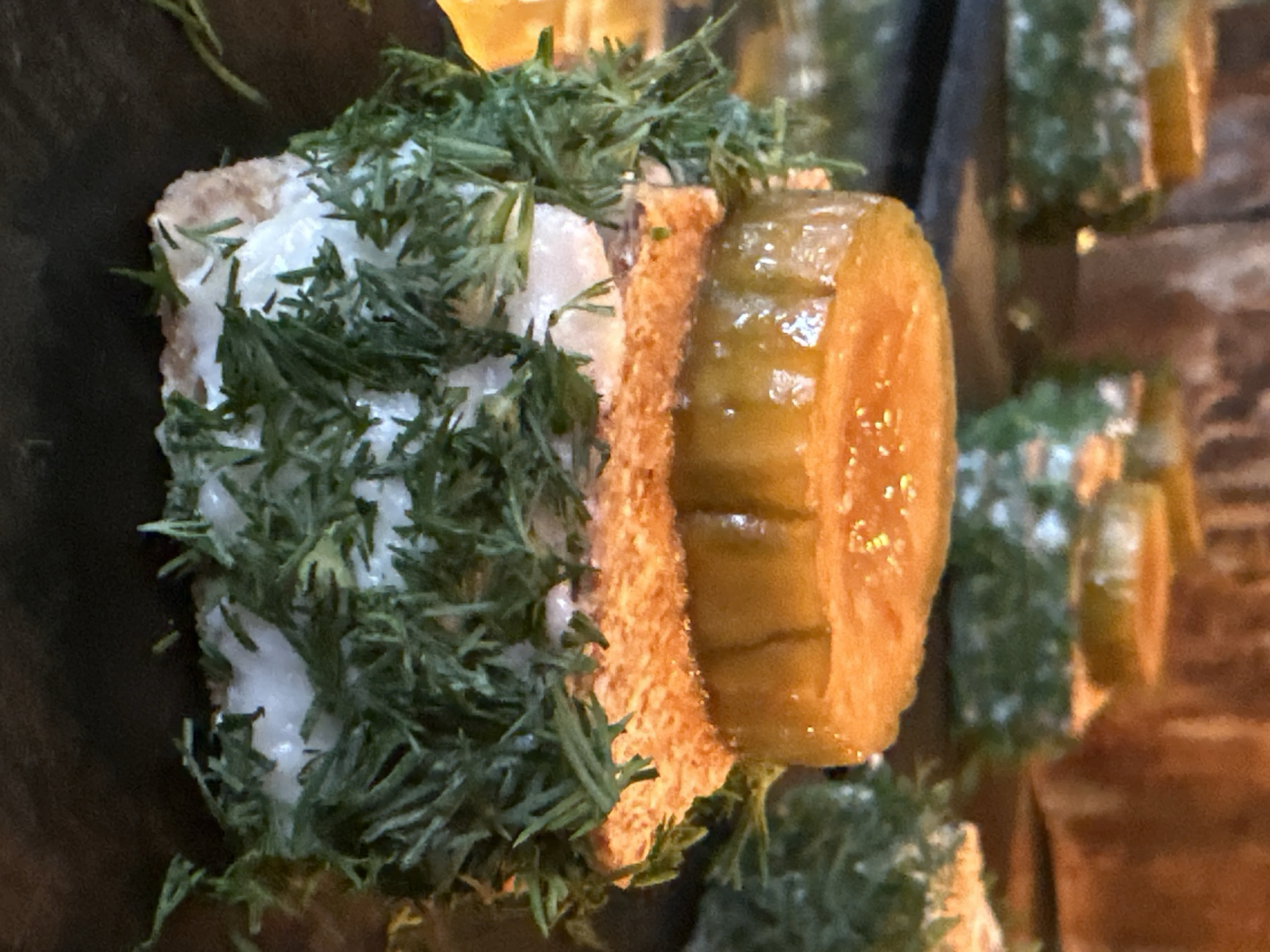 A close-up shot depicts a canapé featuring bread, cream cheese, dill, and a pickle. The canapé is composed of two slices of bread, likely sourdough, sandwiching a layer of cream cheese and topped with a generous amount of fresh dill. A thick, pickled cucumber slice is prominently placed beside the canapé. The image appears to be taken at a food service establishment, as other similar canapés are visible in the background, though blurred. The lighting and ambiance suggest a warm, indoor environment. While the prompt mentions Новоглаголево, Russia, there is no intrinsic visual evidence to confirm that location. The description focuses on the canapé's physical characteristics.