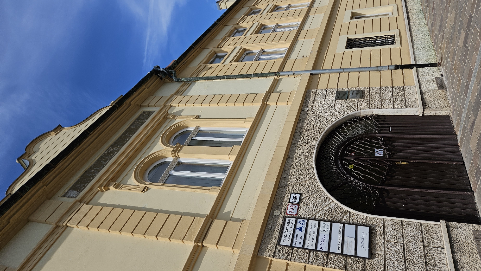 The image shows a low-angle exterior shot of a pale yellow building with light yellow trim, situated on a sunny day under a clear blue sky with wispy white clouds. The building has a pitched roof with a curved gable. Multiple arched windows with white frames are visible on the upper floors. A signpost is mounted on the stone facade near a large, arched, dark brown metal double door. The signpost lists multiple organizations including "KATOLICKY KRUH," "FARIKA KNIZNICA," "LATINSKÝ ZBON," and "KLUB KRESTANSKYCH PEDAGOGOV." Below the address "JARKOVA 77, PREŠOV," the sign also shows "77B 15827." To the right of the entrance, a downspout runs vertically along the building. The foreground features a paved walkway.