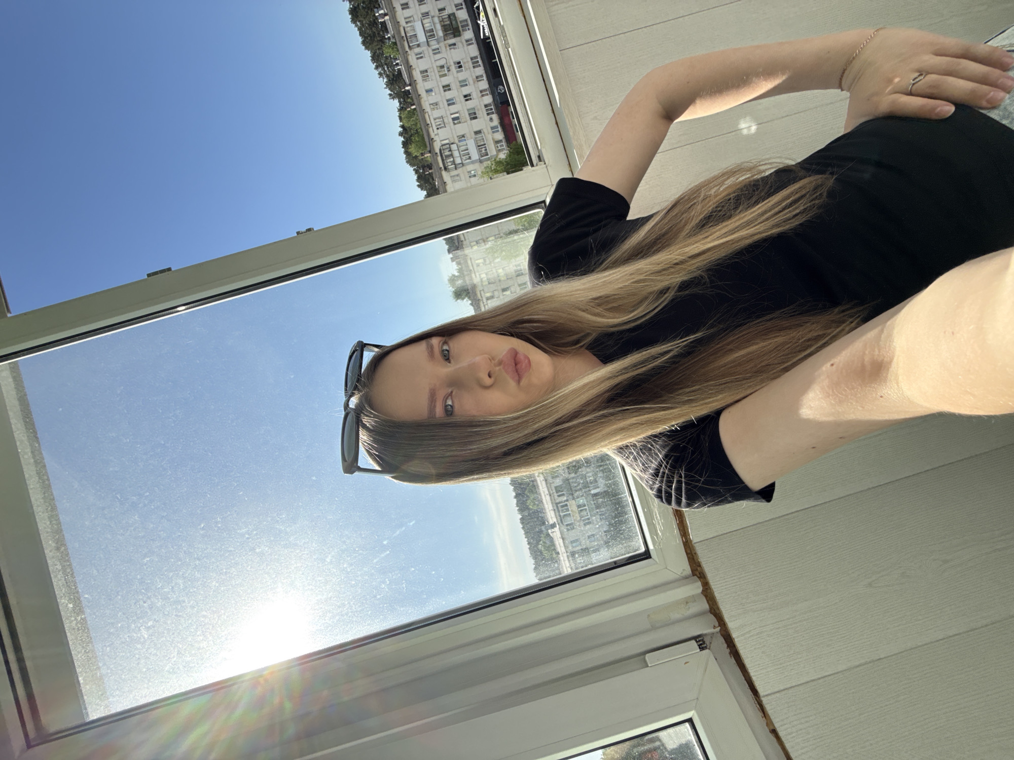 A young woman with long blonde hair is taking a selfie on a balcony. She is wearing a black shirt. Sunglasses are placed on her hair. A ring is visible on her finger. The background shows an apartment building complex in the city of Chaikovsky, Russia, with clear blue skies. The sun is shining brightly through the window.