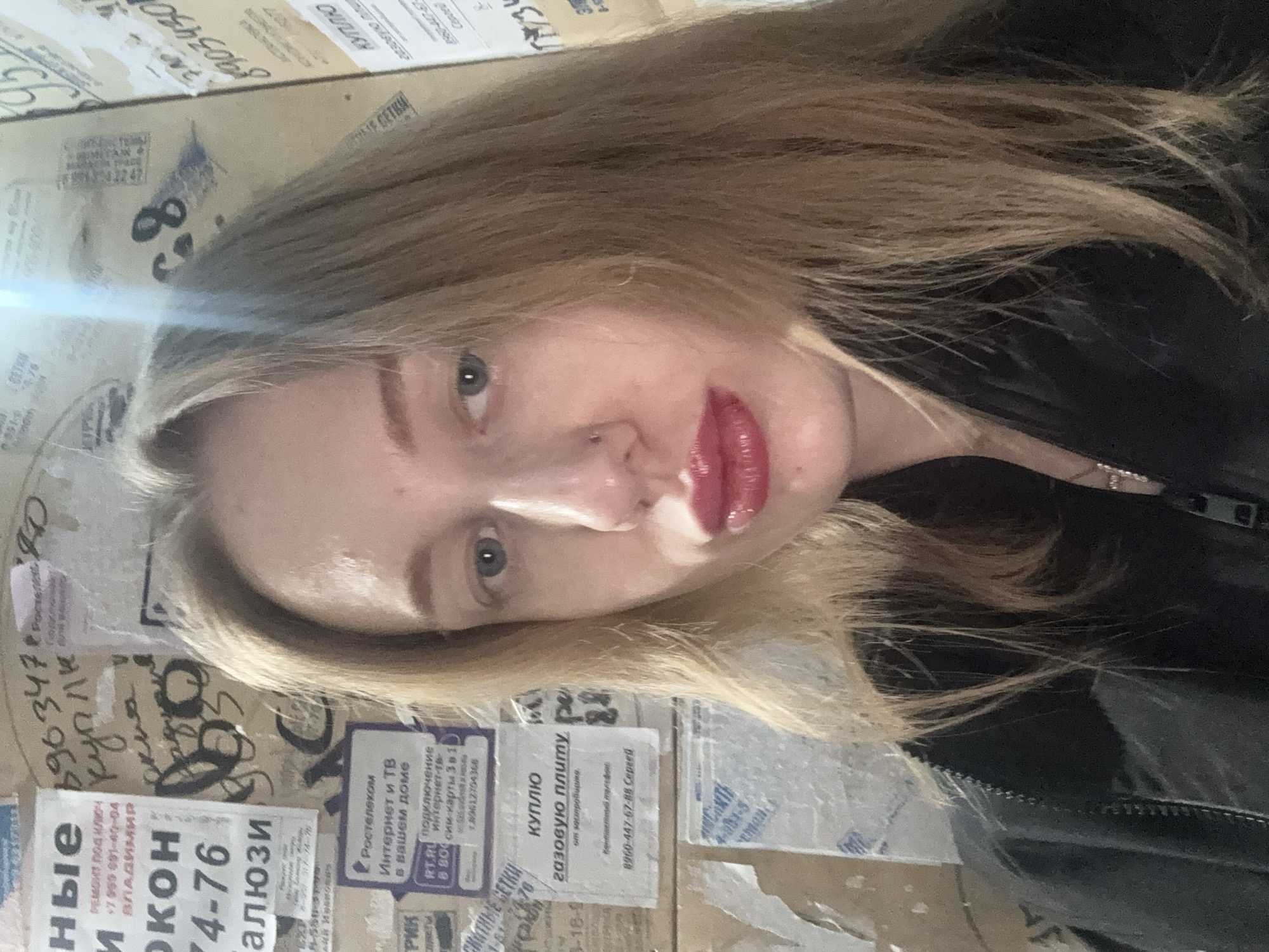 A young woman with blond hair and fair skin is posing for a selfie. She has pink lipstick on. The background appears to be a wall covered with flyers, some of which are advertisements for internet and television services, and a flyer advertising window repair with a phone number starting with +7 999. Some of the flyers are in Russian. Based on the OCR text, the advertisement says it is in Vladimir. The text on the flyer indicates the city of Kuleshovka, Russia.