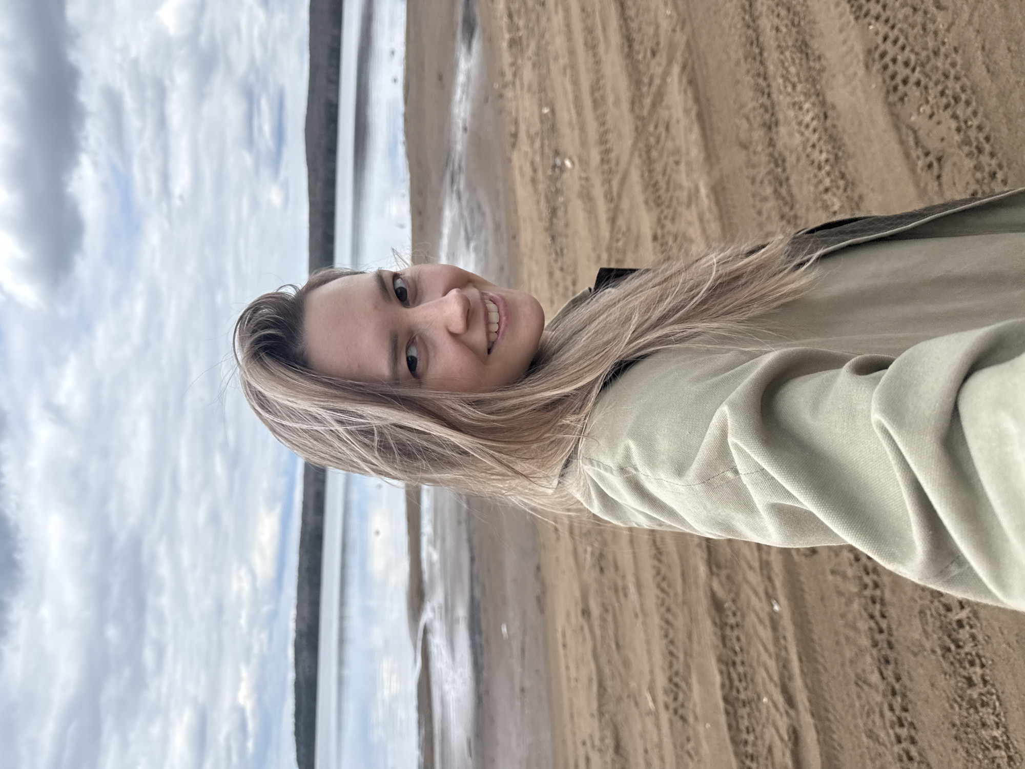 A vibrant outdoor selfie features a smiling young woman, rotated 90 degrees clockwise from a typical upright orientation. The woman, with long, straight blonde hair cascading around her shoulders, is positioned on the right side of the frame. She wears a light olive green jacket with a darker collar peeking out. Her face is turned slightly towards the camera, showcasing a broad, genuine smile with visible teeth, and her light-colored eyes convey happiness and warmth as she looks directly at the viewer.
The background to her left reveals a natural landscape under a cloudy sky. The sky is filled with varying shades of grey clouds, indicating an overcast or partly cloudy daytime. Below the horizon, a calm body of water, likely a lake or the wide Kama River given the Chaikovsky, Russia location context, stretches into the distance, reflecting the muted sky. A dark, narrow band of land is faintly visible on the far side of the water. The immediate foreground and midground comprise a sandy or muddy shoreline, marked with distinct parallel tracks or patterns, possibly from vehicle tires or foot traffic, suggesting a well-traversed area by the water's edge. The scene evokes a tranquil outdoor setting, with the woman's cheerful expression adding a personal and inviting element to the natural backdrop. No text is visible in the image.