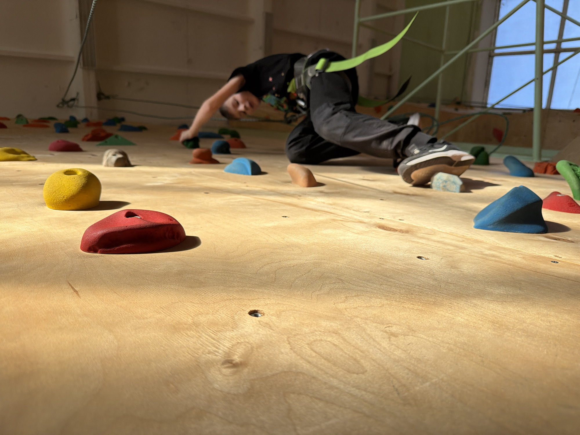 A young person is wearing climbing gear and climbing on an indoor climbing wall in Izhevsk, Russia. The climbing wall is made of plywood and has various colorful climbing holds attached to it. The climber is wearing a black t-shirt, dark pants, and climbing shoes. They are bent over, with one hand on a climbing hold and one foot on another. There is a window in the background, and sunlight is shining into the room.