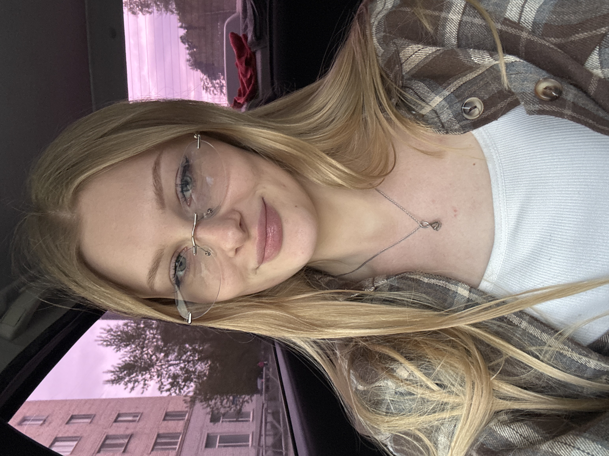 A young, fair-skinned woman with long blonde hair and blue eyes is photographed indoors, likely in a car, as indicated by the window and car interior visible. She is wearing rimless glasses, a white top, and a brown and grey plaid shirt. A silver necklace with an infinity pendant is visible around her neck. The window shows a blurred view of an apartment building and a tree, with the sky appearing pinkish or purple, possibly due to the car's window tint or the time of day (sunset/sunrise). Based on the architectural style of the building visible through the window, and the presence of Russian text in the image context, it is highly probable that the location is Perm, Russia.