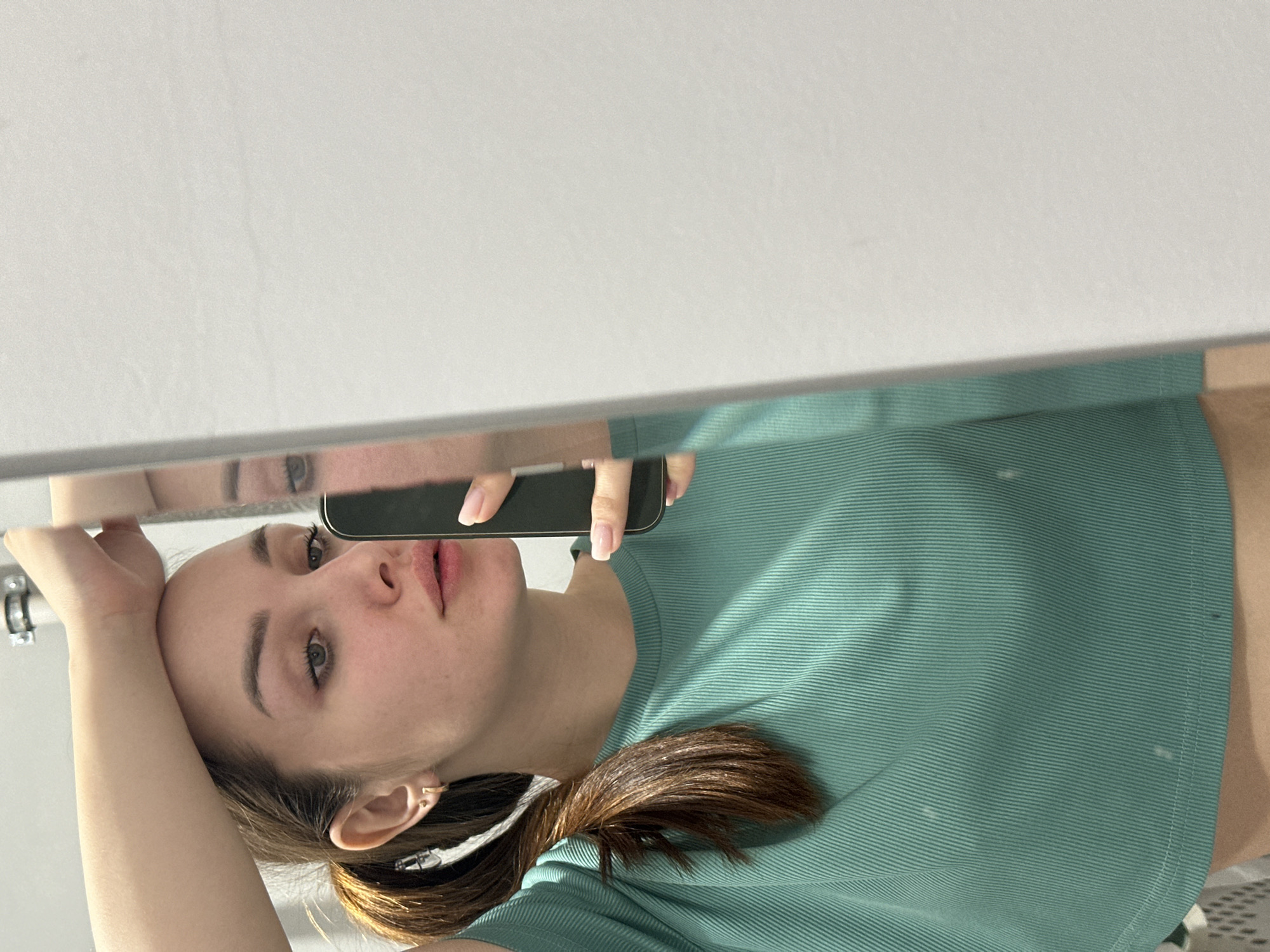 A young woman is captured in a mirror selfie, taken indoors in Chaikovsky, Russia. She is looking directly into the mirror with a neutral expression, her lips slightly parted. Her light brown hair is pulled back in a ponytail, and she is wearing a ribbed, teal-colored short-sleeved top. With her right hand, she holds a black smartphone to take the picture, while her left hand rests casually on her head or forehead. A small, simple earring is visible in her right ear. The background reflected in the mirror is mostly a plain, light-colored wall or ceiling, indicating a bright indoor setting. There is no visible text in the image. The overall impression is one of a candid, casual moment.