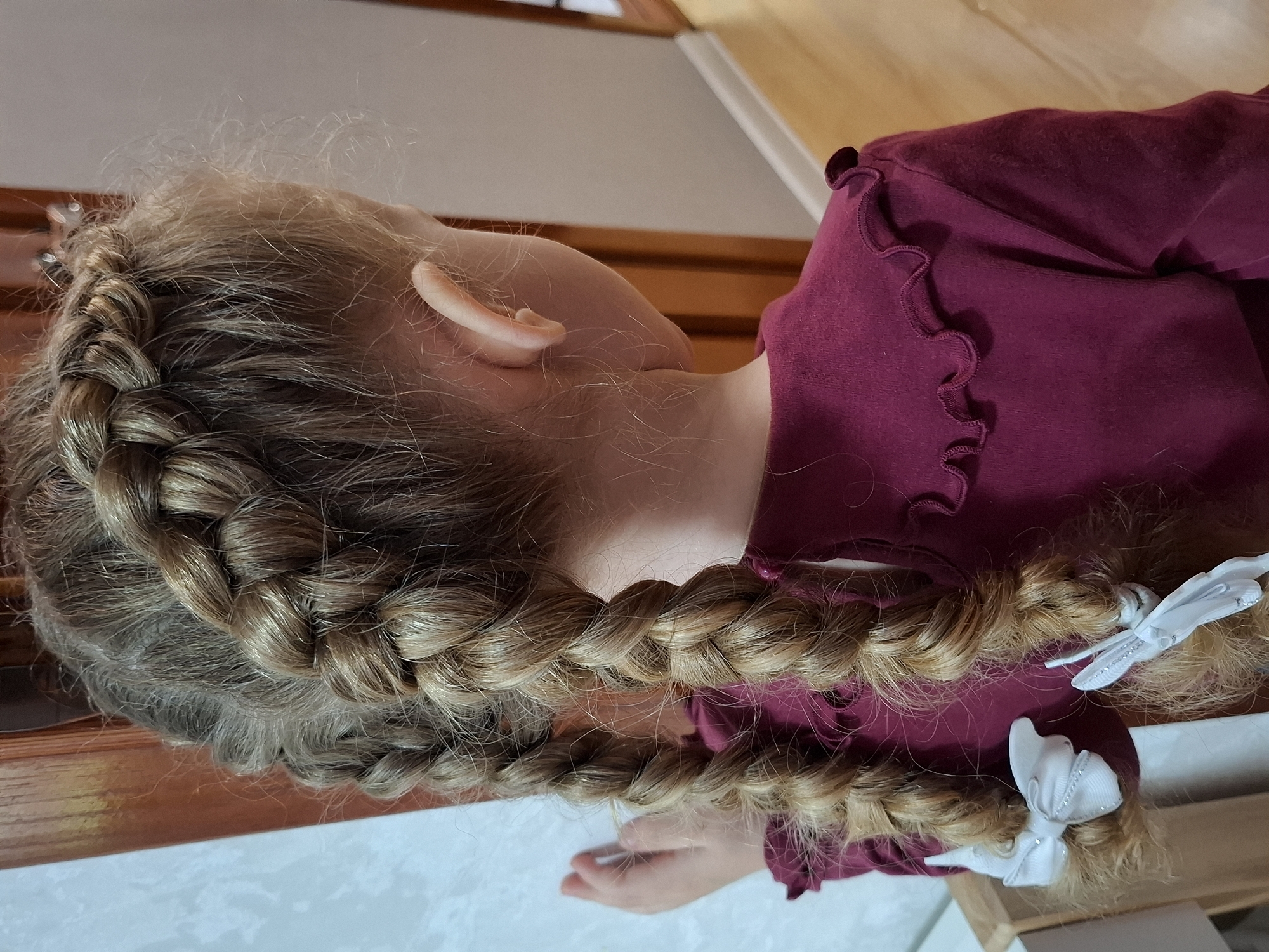 A young person, likely a child, is depicted from a rear-facing perspective in an indoor setting in Chaikovsky, Russia. Their light blonde or light brown hair is intricately styled into two French braids that extend down their back. Fine, wispy flyaway hairs are visible around the scalp and hairline, giving the appearance of soft, slightly unkempt strands despite the neat braids.
The subject is wearing a maroon or burgundy long-sleeved top made from a soft, textured fabric, possibly velour or fleece. The top features a decorative ruffled trim along the neckline or shoulder seam. Each braid is secured at its end with a white hair bow. One of these bows is further adorned with a delicate white, possibly beaded, insect-shaped charm, resembling a dragonfly or butterfly. A small, pale ear is visible on the side of the head, and a partial view of a child's hand is seen in the lower right portion of the frame.
The background shows elements of an indoor environment, including warm-toned wooden surfaces, possibly furniture or a floor, and lighter walls or ceilings. The lighting is soft and diffused, characteristic of indoor daytime conditions. No specific activity is taking place, and emotions are not discernible from this rear view. There is no visible text in the image.