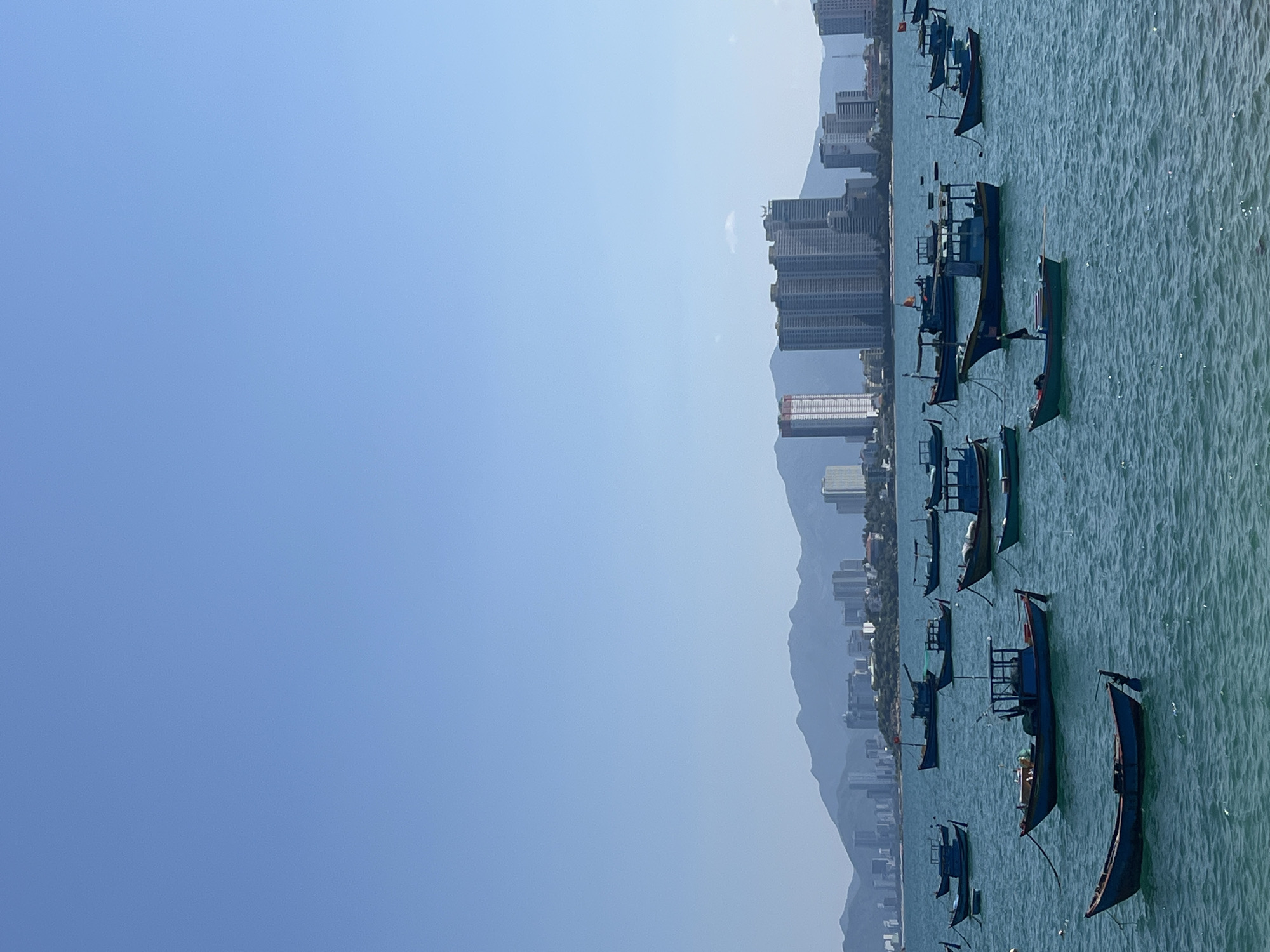 The image displays a panoramic daytime view of Nha Trang, Vietnam. The upper portion consists of a clear, unblemished blue sky. In the background, a range of hazy, distant mountains is visible. Below the mountain range, the city's skyline extends along the coastline, characterized by numerous multi-story buildings and high-rise structures, presenting a mix of white, grey, and red-accented facades. The midground and foreground feature a body of turquoise-blue water with a rippled surface, indicating gentle wave activity. Distributed across this water are an estimated 20 to 30 traditional Vietnamese wooden fishing boats. These boats are predominantly painted blue, with some featuring red, white, or natural wood elements, and many include small cabins or shelters. All visible boats are stationary, suggesting they are anchored or moored. No individuals are discernible within the frame.