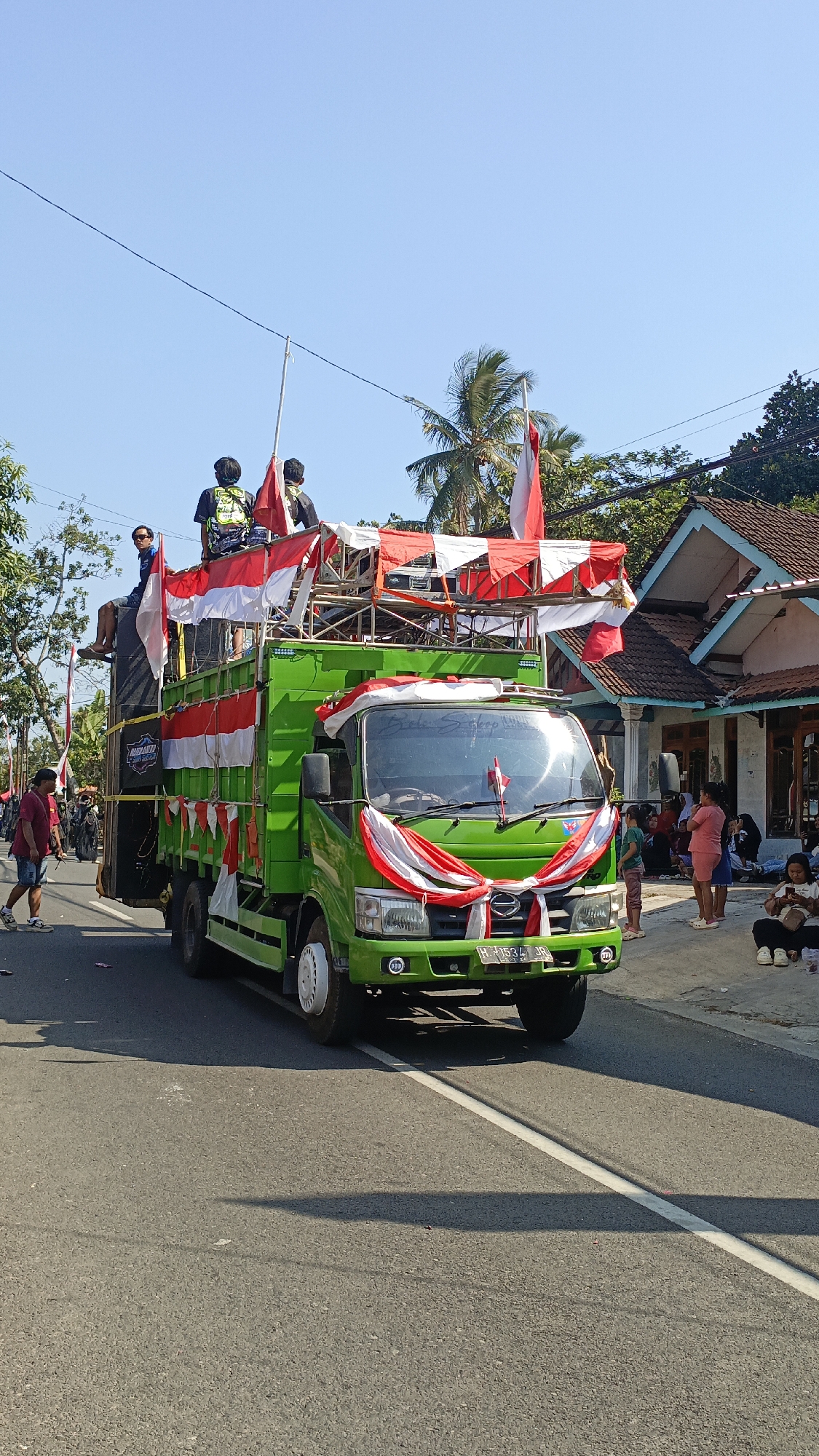 A green truck, adorned with red and white Indonesian flags and fabric streamers, proceeds down a paved road in Krajan Lor, Indonesia. Two individuals are seated on top of the truck's structure, while another person stands to their left. The truck is equipped with large speakers on its left side. On the right side of the road, several people are gathered, some seated and watching the truck. Houses with tile roofs are visible in the background, along with trees and a clear blue sky. The scene appears to be a parade or celebration.