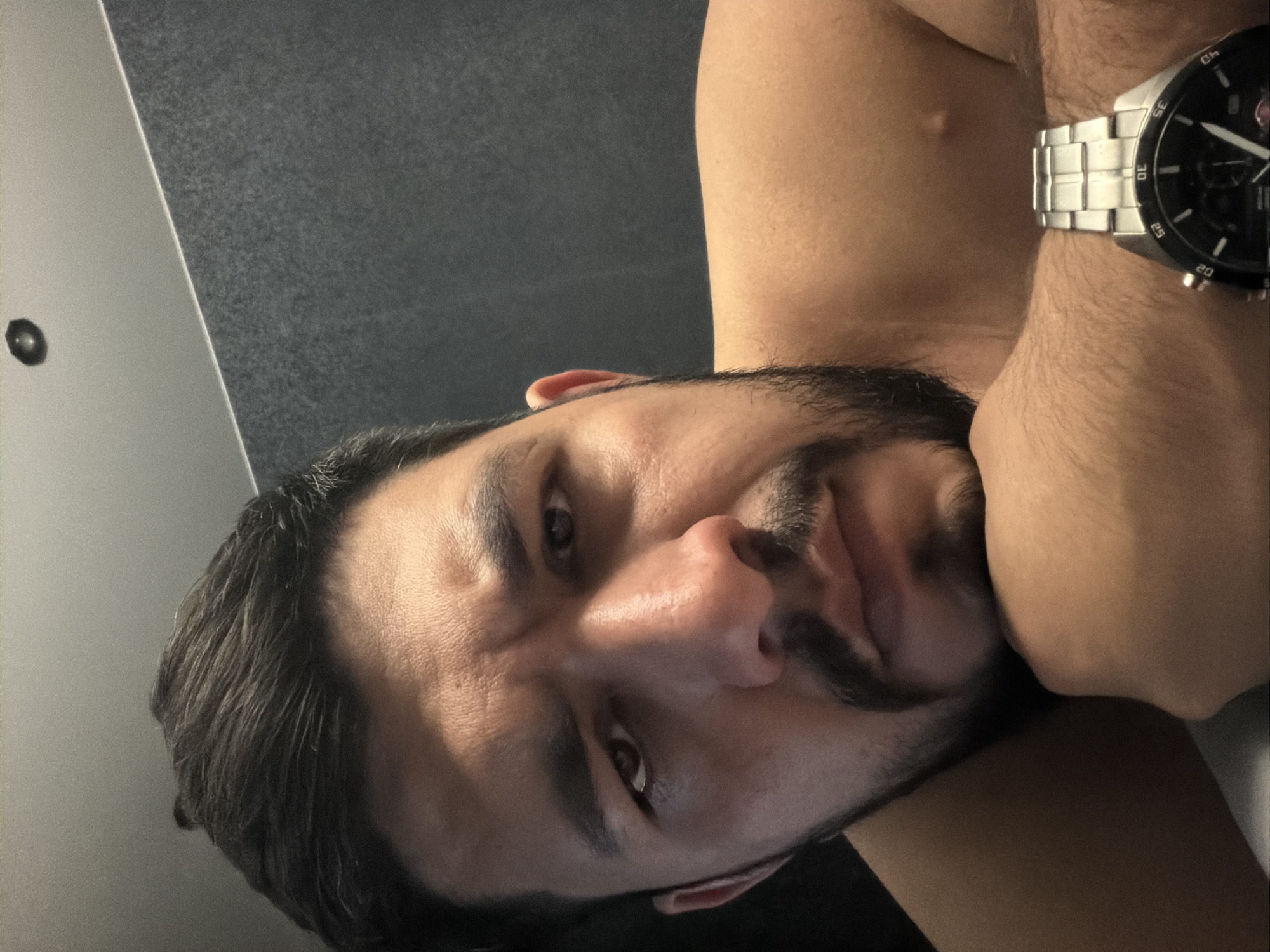 A close-up, selfie-style shot depicts a man with dark hair and a beard. He has an intense gaze directed at the camera. He is wearing a silver wristwatch. The background features a wall, possibly indoors. There is no information present in the image to determine if the image was taken in Krasnoyarsk, Russia or to provide any other details concerning location.