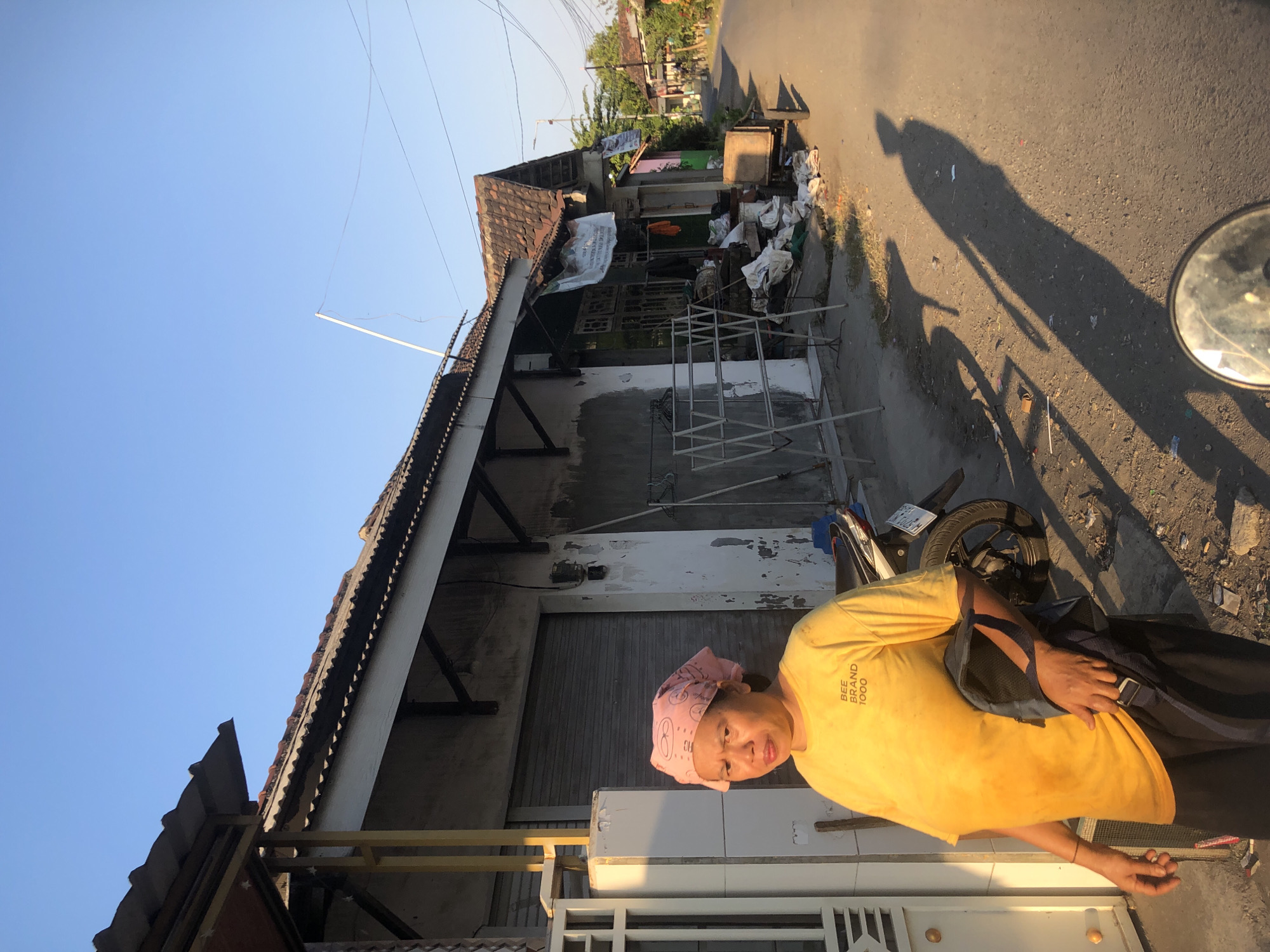 In the city of Kalidawir, Indonesia, a woman wearing a pink bandana and a yellow t-shirt stands by the roadside. The t-shirt has the text "BEE BRAND 1000" printed on it. She is holding a dark bag over her shoulder. The background shows a building with a tiled roof and a closed metal shutter. In front of the building, there is a clothes drying rack and various items piled on the ground. A motorcycle is parked behind the woman. The scene appears to be a street in a residential or commercial area, with buildings and trees visible along the road. The lighting suggests it is either early morning or late afternoon, with long shadows cast on the ground.