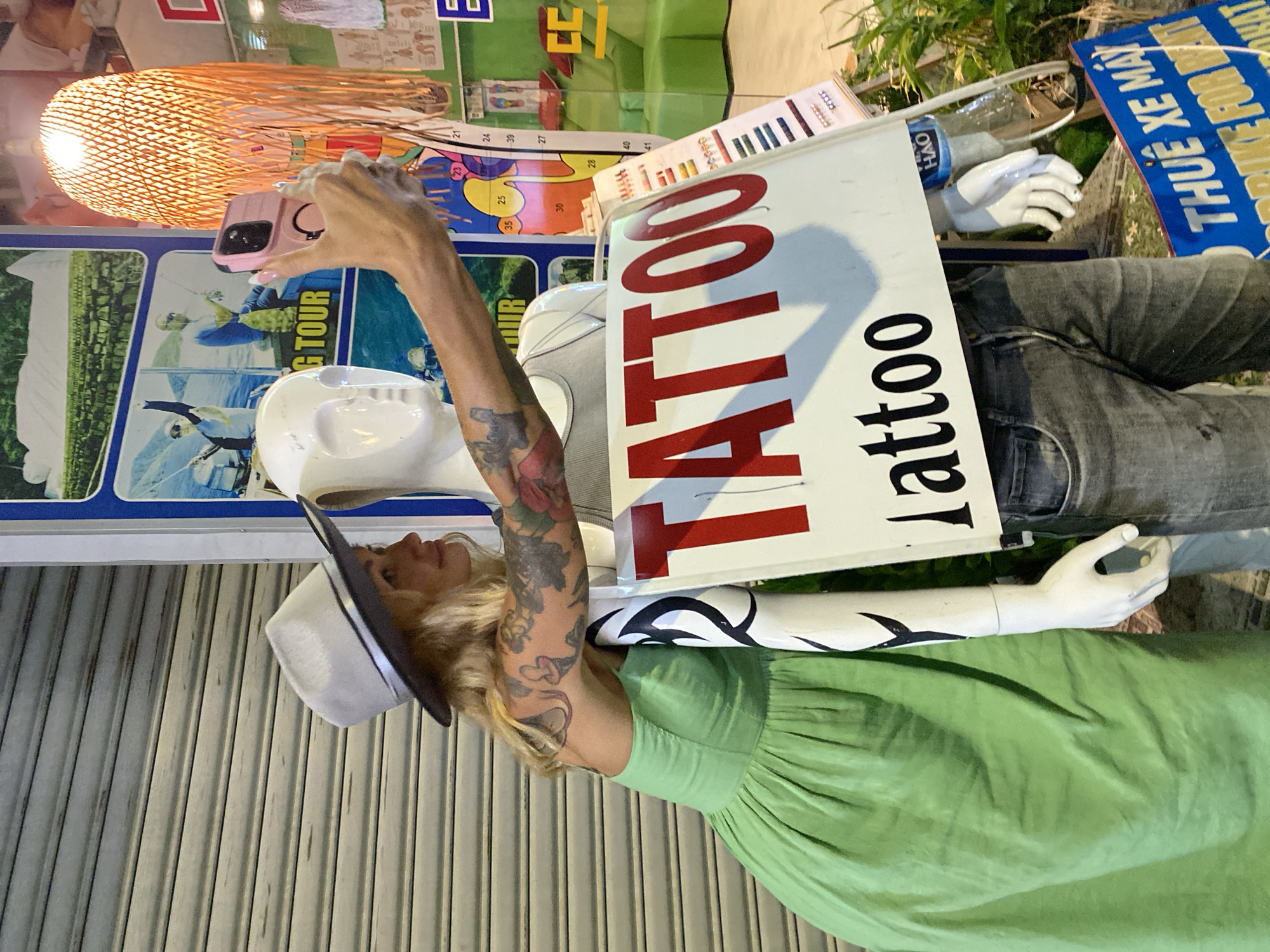 A woman in a light green dress and a grey fedora is taking a selfie with a mannequin that has a sign reading "TATTOO" and "l attoo" on it. The woman has tattoos on her arms. She is holding a pink smartphone in her right hand. The mannequin is wearing grey jeans and a grey tank top. Behind them, a metal garage door is visible. To the right, there is a poster with images of people fishing and text that appears to be in Vietnamese, along with a brightly lit orange lampshade. Further to the right, a window shows a green sofa inside. A blue sign with Vietnamese text, including "THUE XE MAY" and "BIKE FOR RENT", is visible at the bottom of the frame. The scene appears to be an outdoor setting in Phu Thuy Ward, Vietnam, likely on a street.