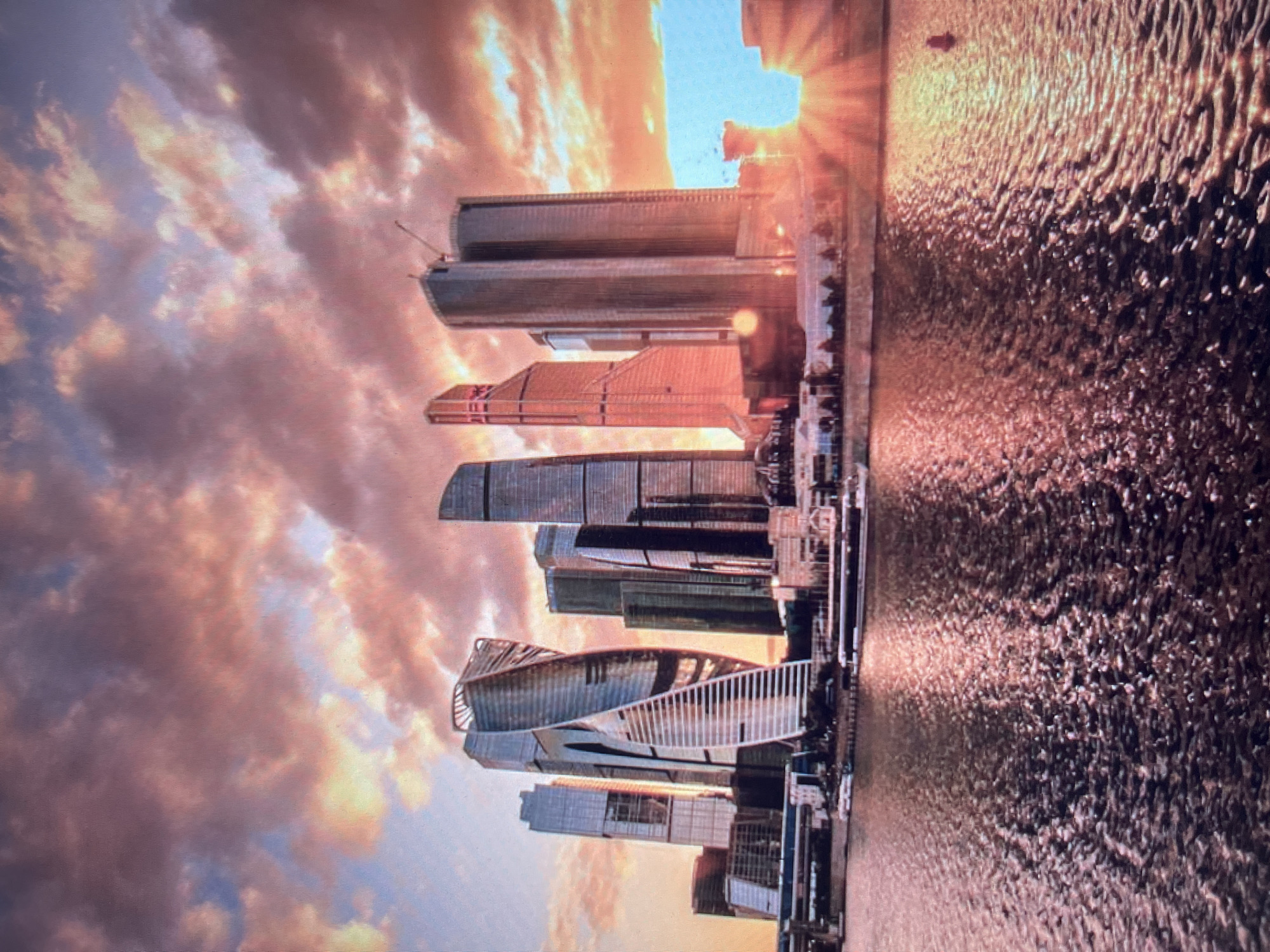 The image depicts a panoramic view of the Moscow City skyline in Khimki, Russia, at sunset. The sky is filled with dramatic clouds tinged with pink, orange, and purple hues. Several modern skyscrapers, characterized by their sleek, geometric designs and glass facades, dominate the scene. The most prominent structure is a uniquely twisted skyscraper, its form spiraling upwards. In the foreground, a body of water, likely the Moskva River, reflects the warm colors of the sunset and the city lights. The water surface is rippled, creating a shimmering effect. The overall impression is one of urban grandeur bathed in the warm glow of the setting sun, with a sense of peacefulness conveyed by the tranquil water. No people or specific actions are discernible in the image.