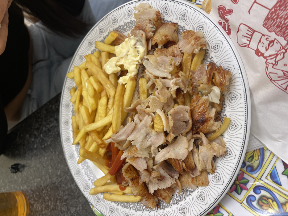 This close-up image captures a large, casual meal served on a plate in an indoor setting, likely in Sondrio, Italy. The central subject is a generous portion of food, predominantly comprising golden-brown french fries and numerous strips of shaved meat, characteristic of döner kebab (appearing to be chicken or turkey). A dollop of creamy white sauce, possibly mayonnaise or garlic sauce, is visible on a pile of fries, with traces of red ketchup also present beneath some of the fries.
The food is presented on a white, circular plate featuring a distinct black and white patterned border composed of repeating heart-like or teardrop motifs. The plate rests on a dark, possibly granite or marble, countertop or table surface. To the right, a vibrant section of a table or placemat with colorful, geometric tile patterns adds a contrasting visual element.
A person is partially visible in the upper left of the frame, showing a dark sleeve or garment, part of a hand with a ring, and dark hair, suggesting they are seated at the table. In the background, a hint of light blue clothing indicates the presence of another individual. At the bottom left, a golden-colored glass, possibly containing a beverage, is partially visible.
To the right of the plate, a crumpled white paper bag or wrapper is visible. It features red text, with the letters "ER" clearly discernible, and a stylized illustration of a chef's head and hat. This packaging suggests the meal may be from a takeaway or a casual eatery. The scene depicts a typical mealtime, such as lunch or dinner, in a relaxed environment.