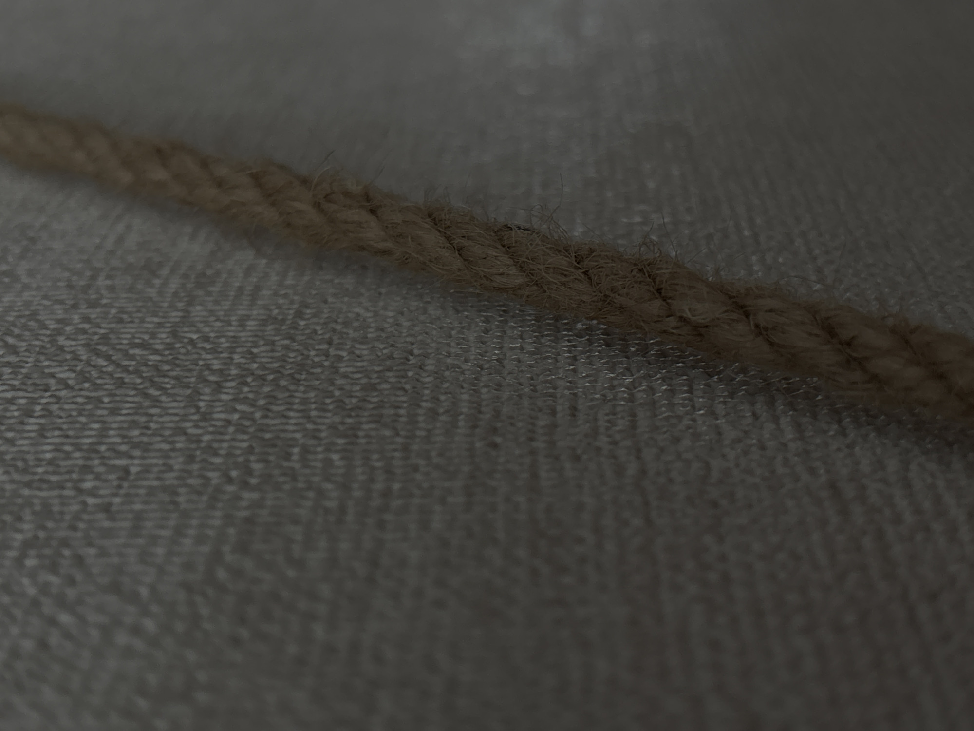 The image displays a close-up, angled view of a thick, woven rope against a textured, neutral-colored surface. The rope, a light brown or tan color, appears to be made of natural fibers, possibly jute or hemp, and shows signs of wear with some frayed strands. The background surface has a subtle pattern of small, raised dots or a tightly woven weave, creating a slightly rough texture. The lighting is dim, casting soft shadows and highlighting the textures of both the rope and the background. The image is abstract and lacks any discernible context of a specific location within Saint Petersburg, Russia.